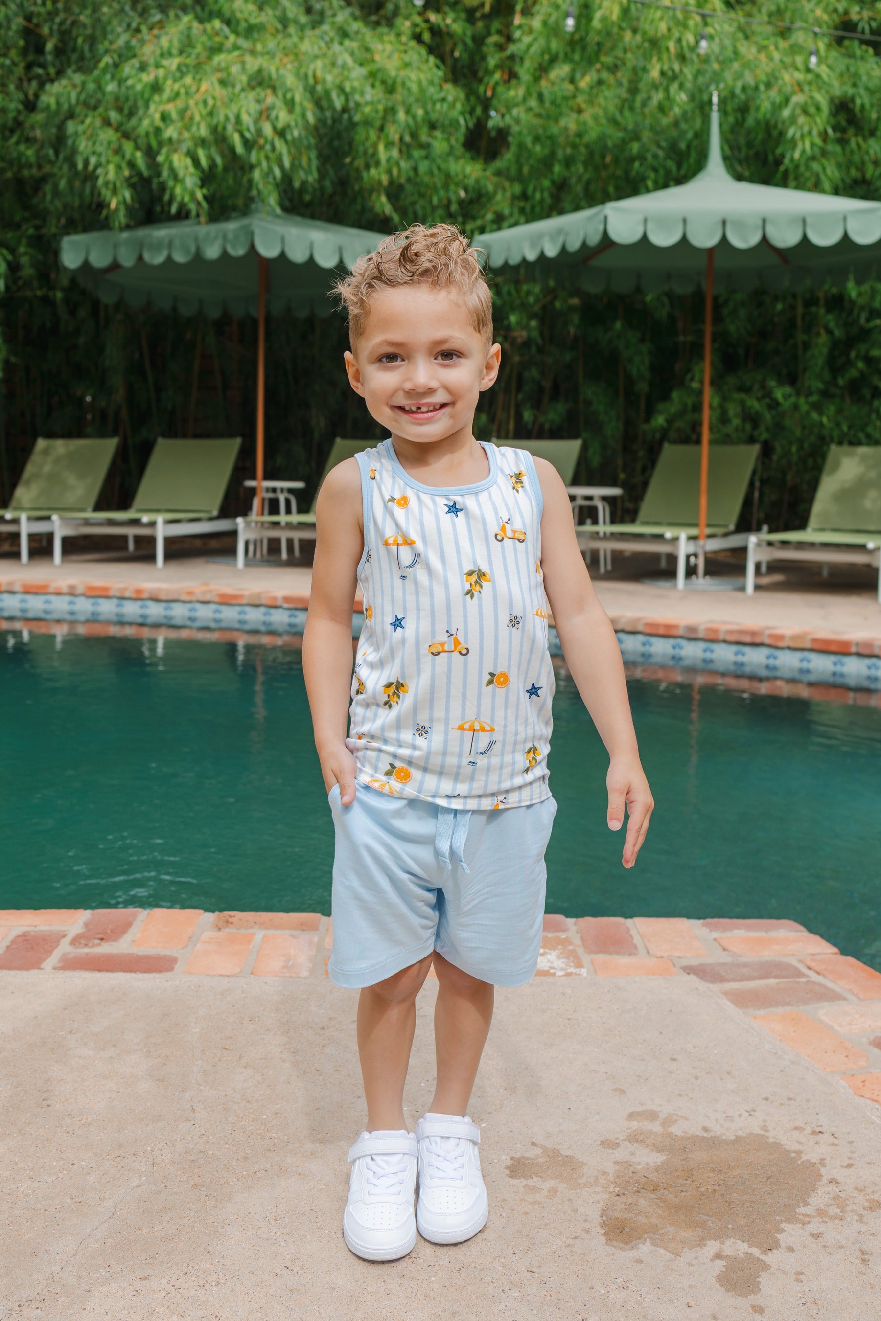 Young boy wearing a tank top with amalfi coast inspired print and blue shorts by a pool