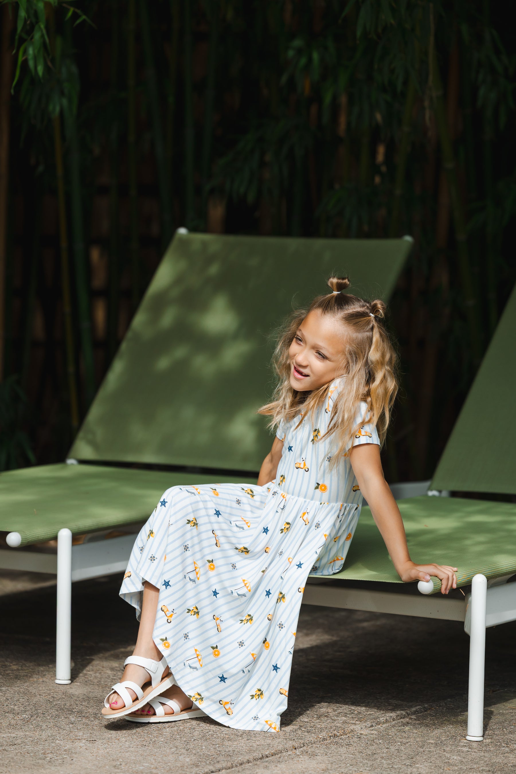 Young girl in a short sleeve dress with blue stripes and amalfi coast inspired icons sitting on a green lounge chair outdoors.