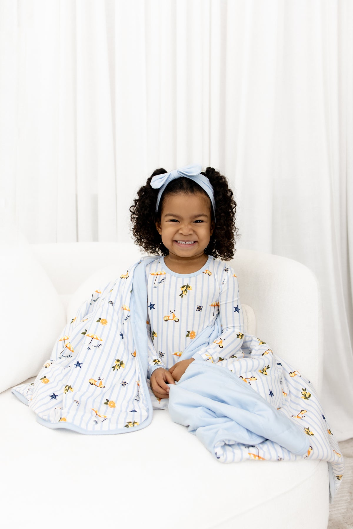 Smiling girl sitting on a white couch with the Toddler Blanket in Amalfi Coast 1.0 wrapped around her wearing matching long sleeve pajamas