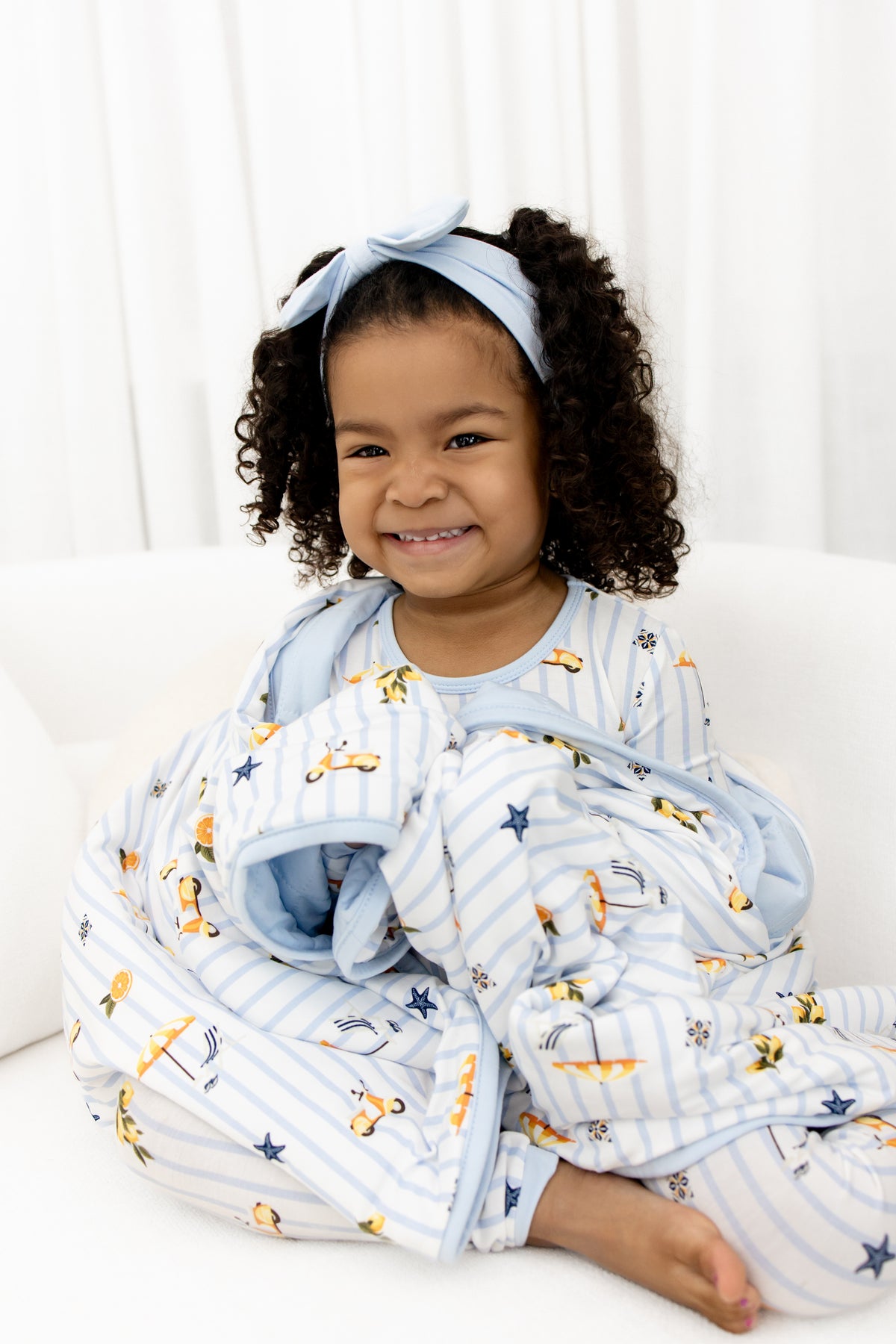 Close up of young girl sitting on a white couch with the Toddler Blanket in Amalfi Coast 1.0 wrapped around her wearing matching long sleeve pajamas
