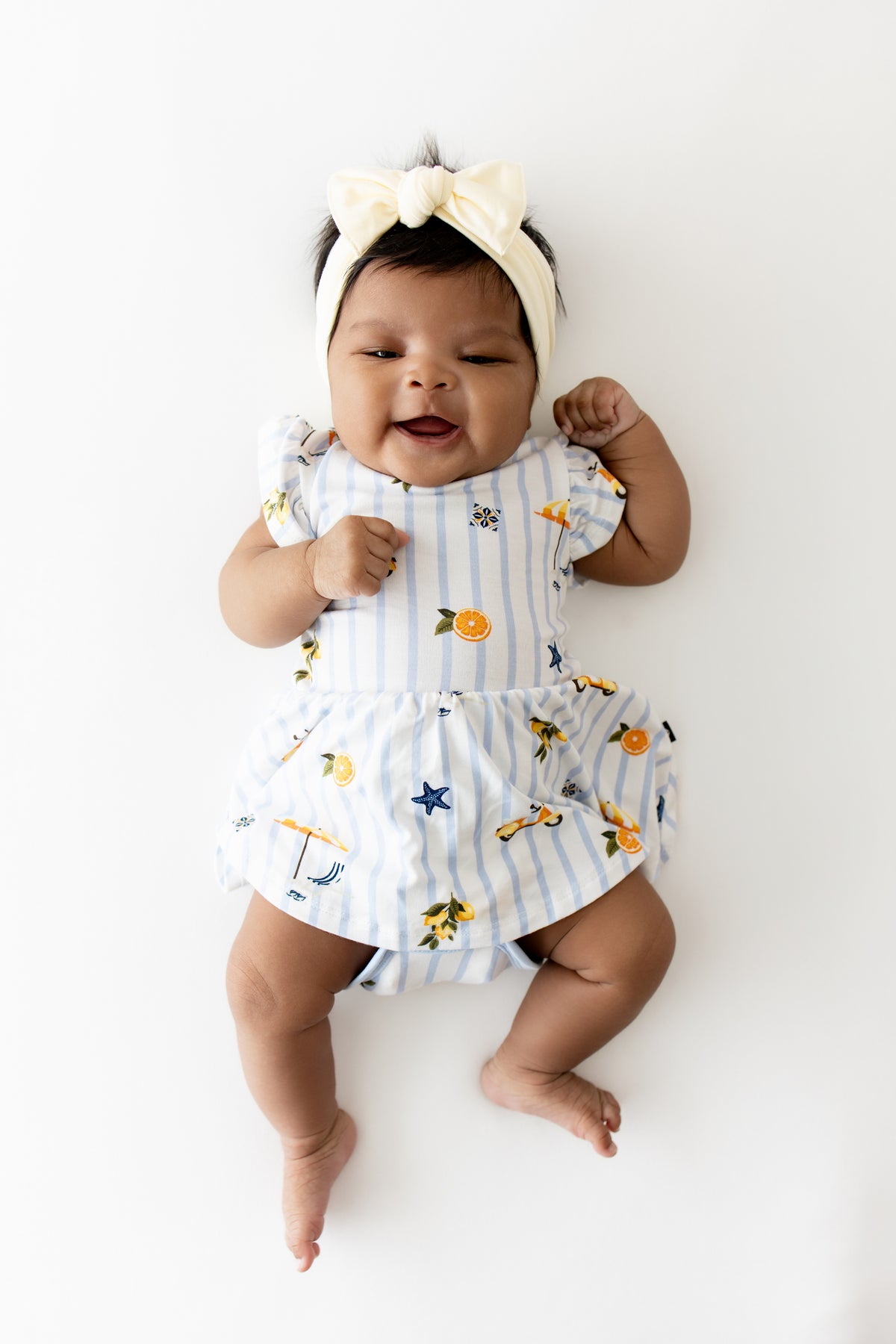 Infant laying on a white surface wearing the Twirl Bodysuit Dress in Amalfi Coast with a light yelllow bow