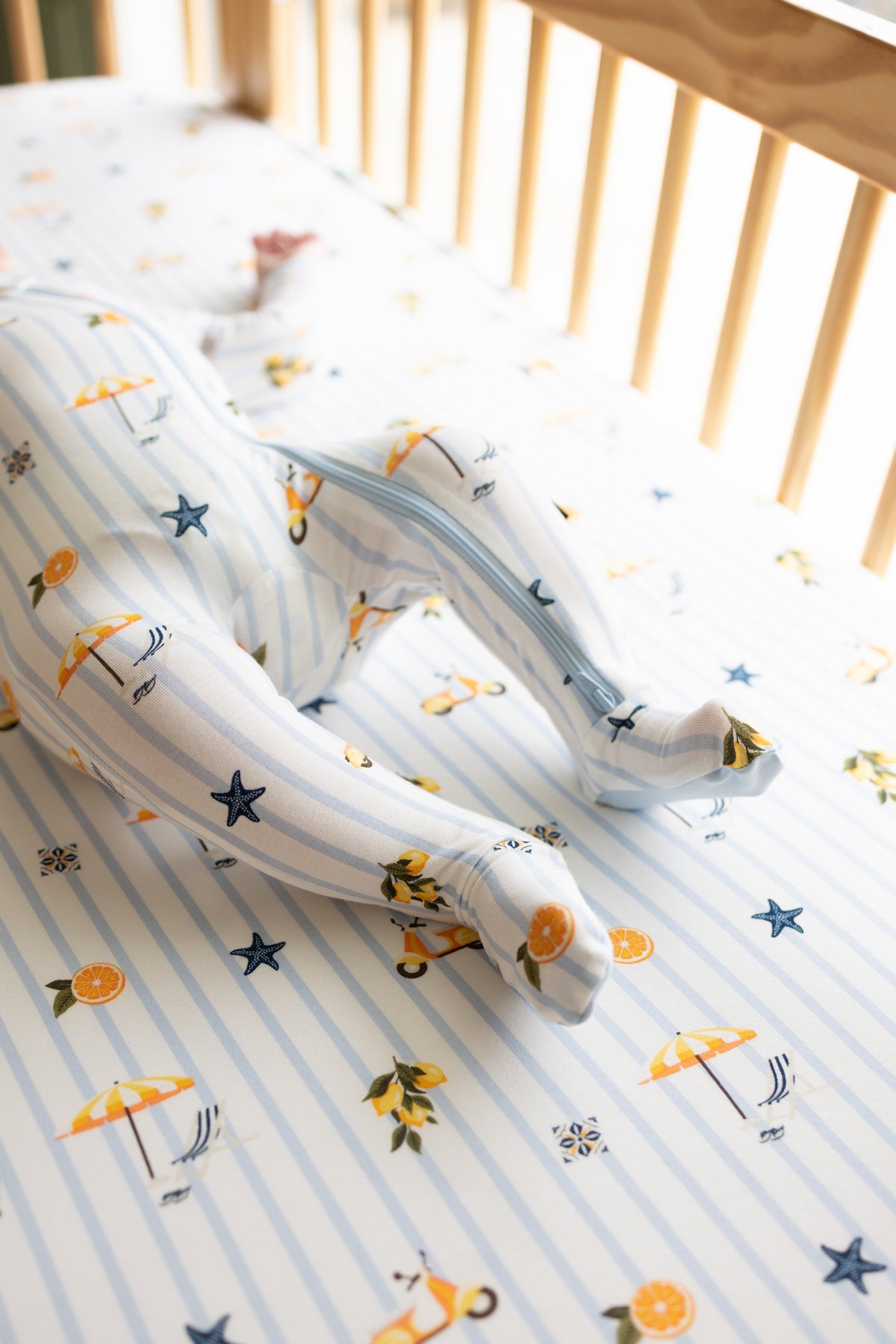Infant laying in a crib on a Crib Sheet in Amalfi Coast wearing a matching zippered footie
