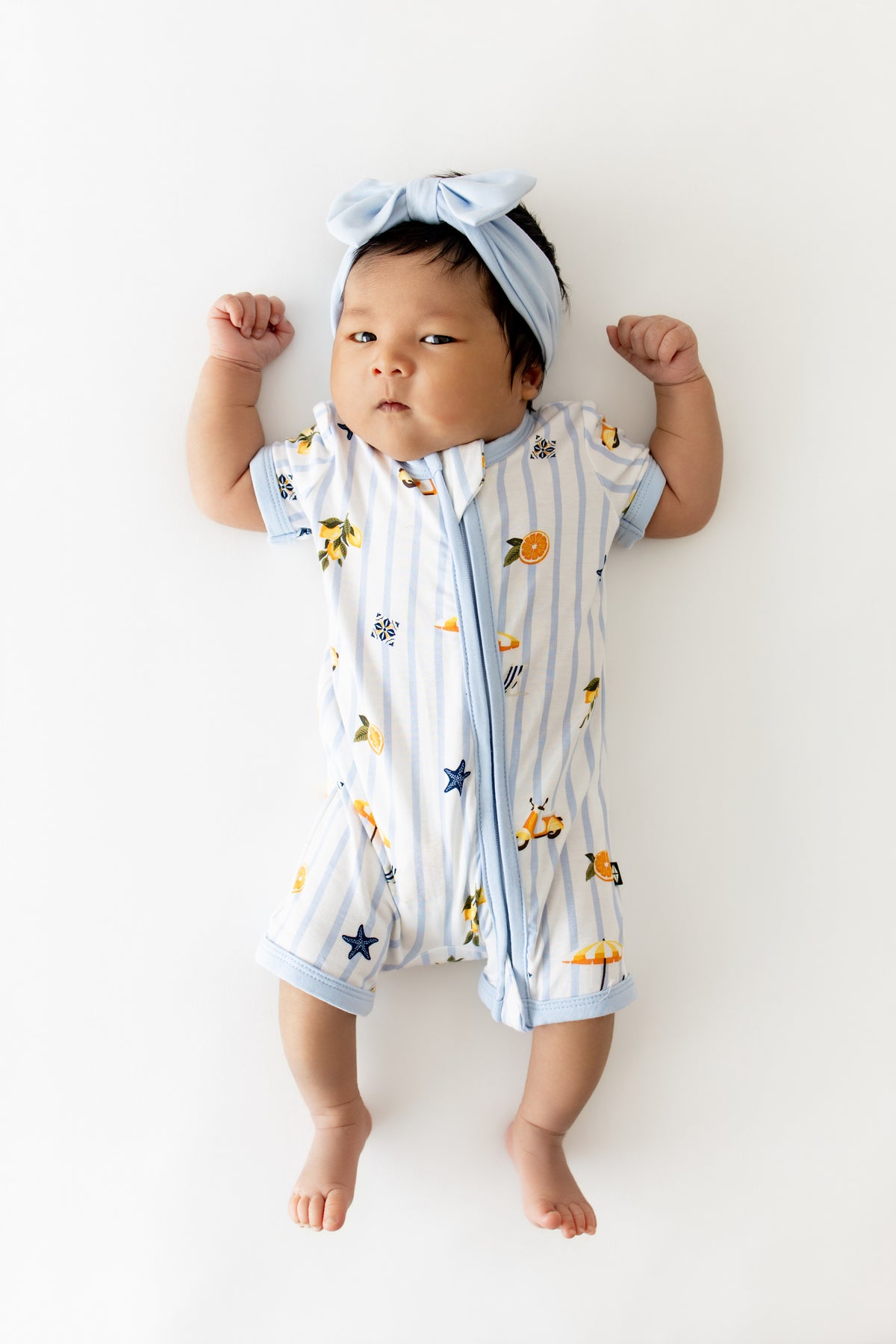 Infant laying on a white surface wearing the Zipper Shortall Romper in Amalfi Coast with a light blue bow