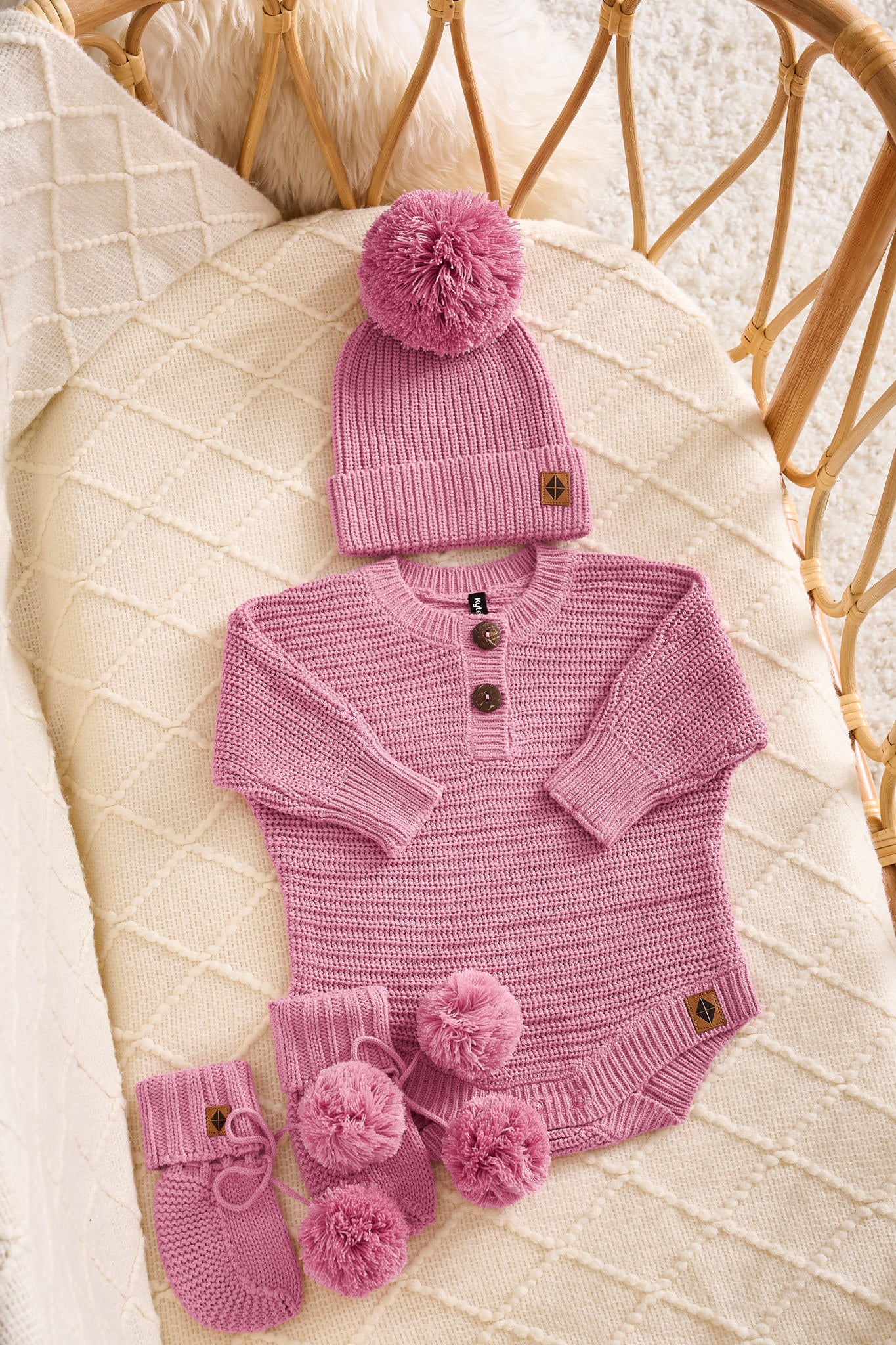 A baby knit outfit set in dusty pink, including a long-sleeve henley bodysuit with three buttons, a matching beanie with a pom-pom, and booties with pom-poms, arranged on a cream quilted blanket in a wicker basket.