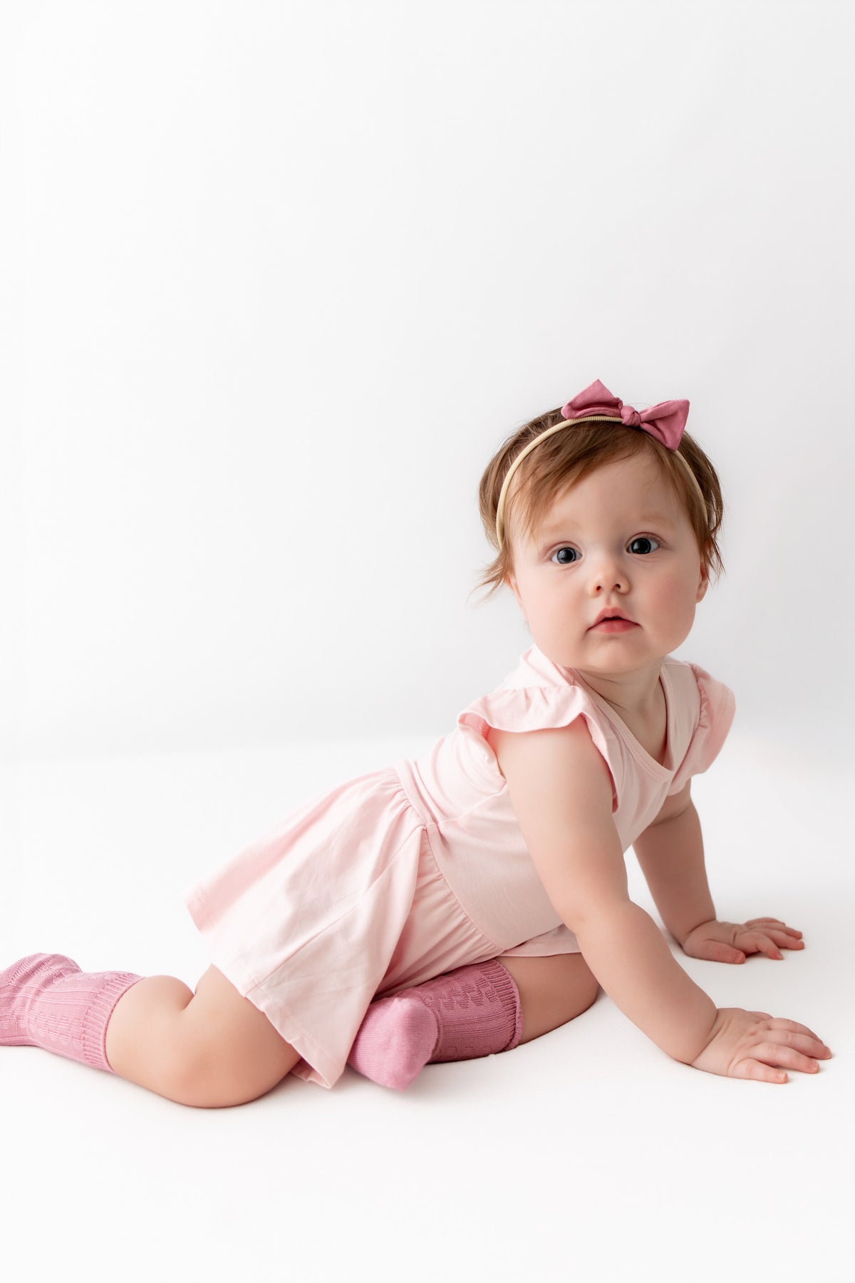 A baby girl crawling on a white studio floor, wearing a light pink ruffle-sleeve dress, pink socks, a pink bow headband, and looking directly at the camera.