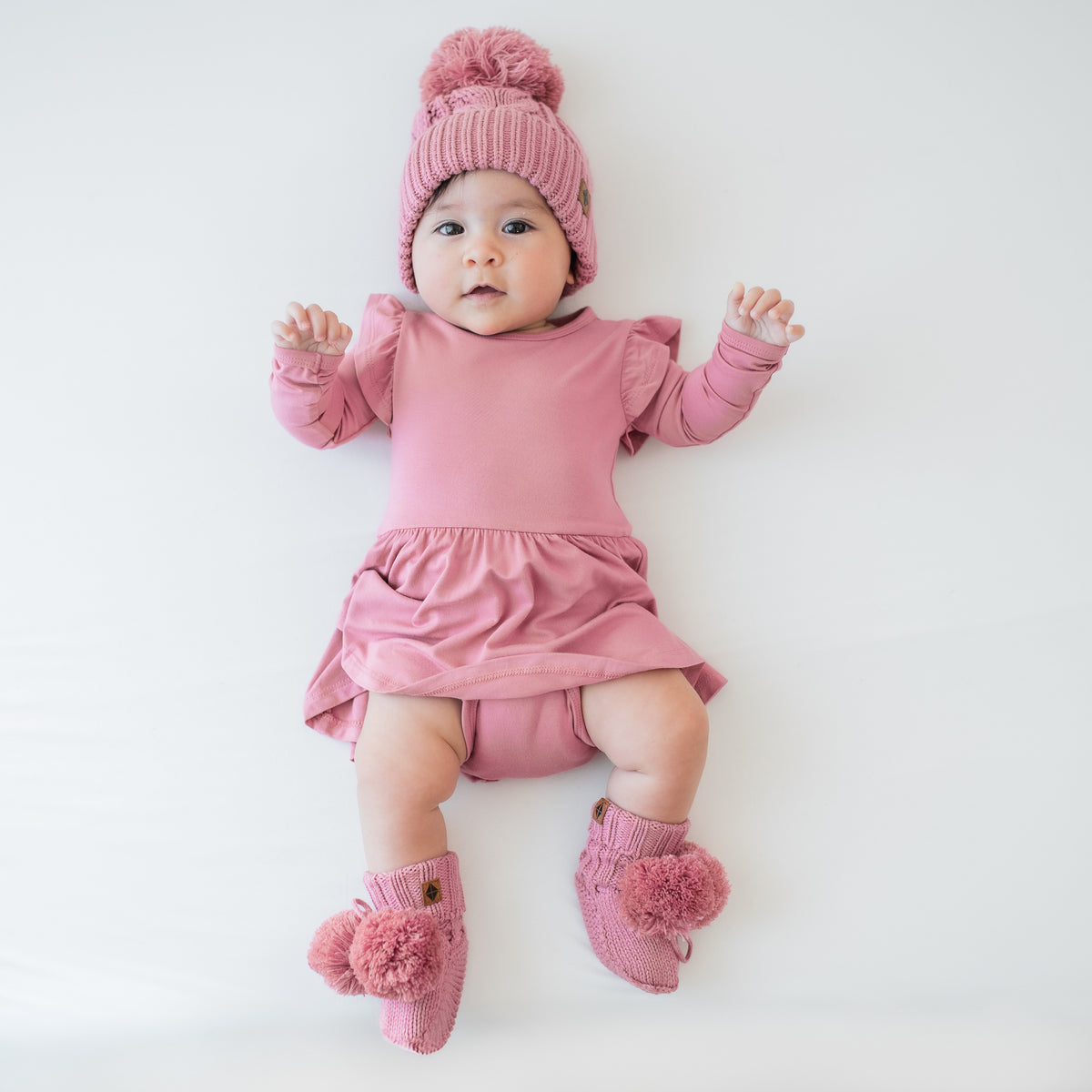 Infant laying down wearing the Knit Booties in Apple Blossom paired with matching long sleeve twirl bodysuit dress and a cable knit pom beanie