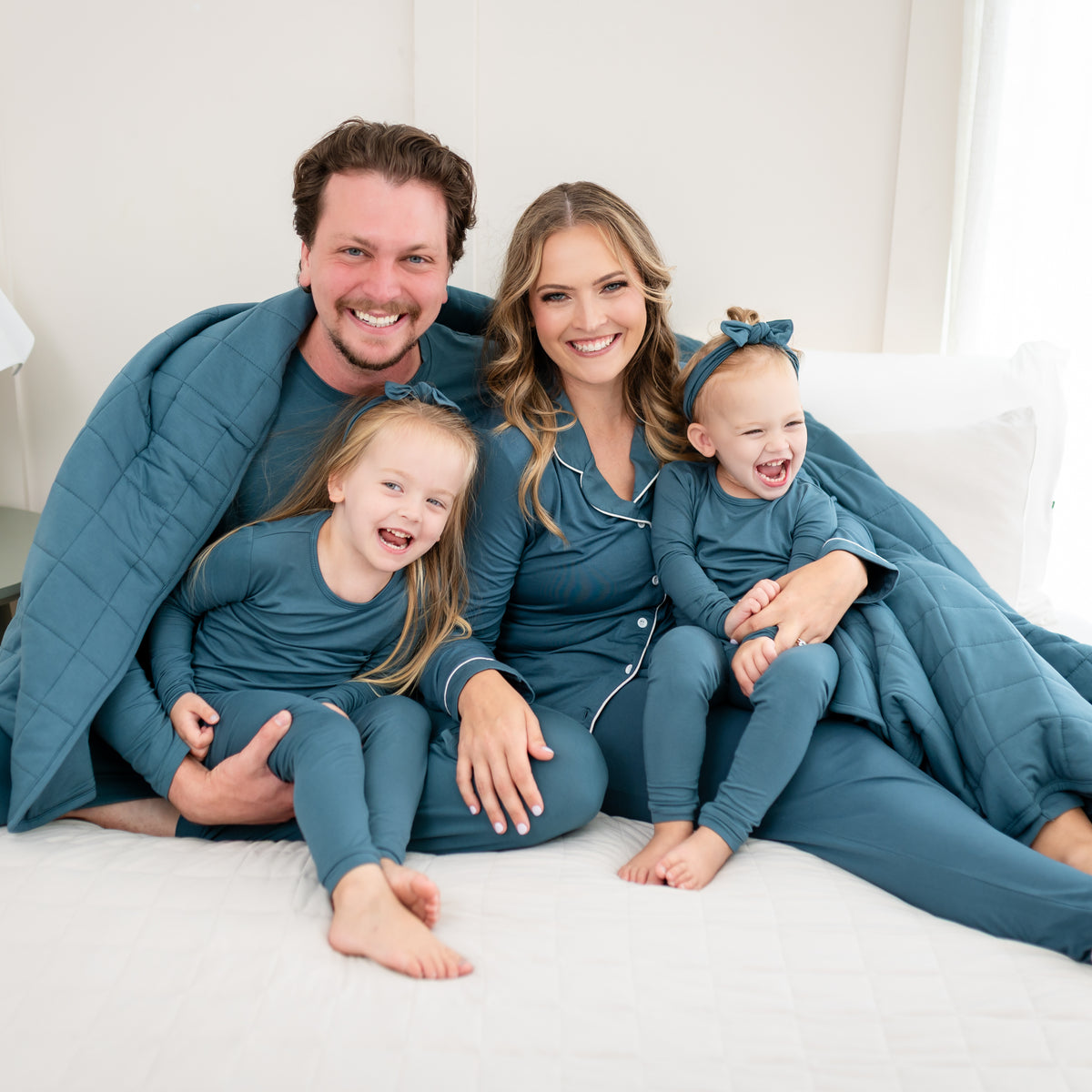Family matching in Atlantic Pajamas sitting on a bed with an Adult Quilted Blanket in Atlantic 2.5 draped over the parents
