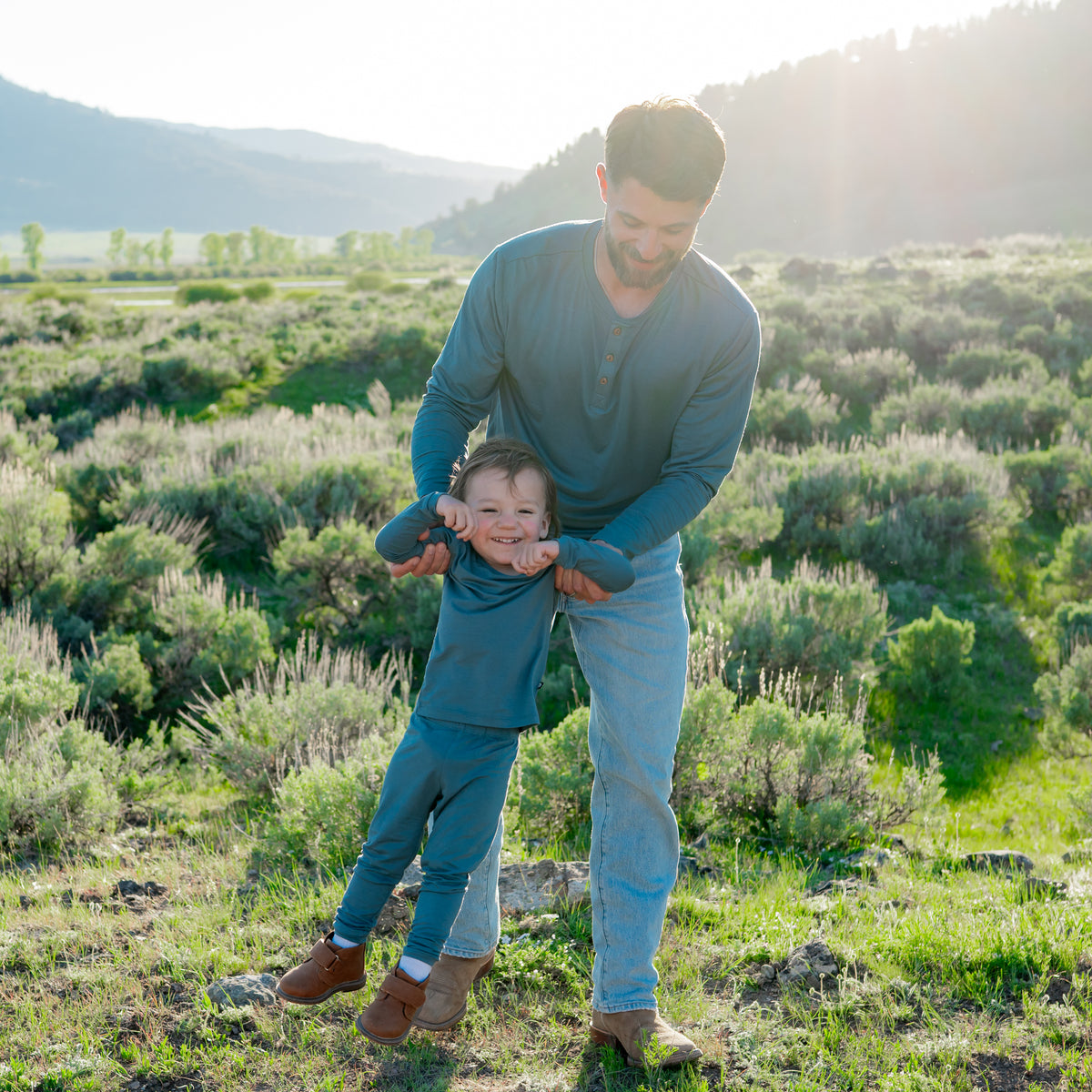 Father holding child under child's arms outside in a field. Father wearing Men's Long Sleeve Henley Tee in Atlantic and child is wearing the Long Sleeve Pajamas in Atlantic