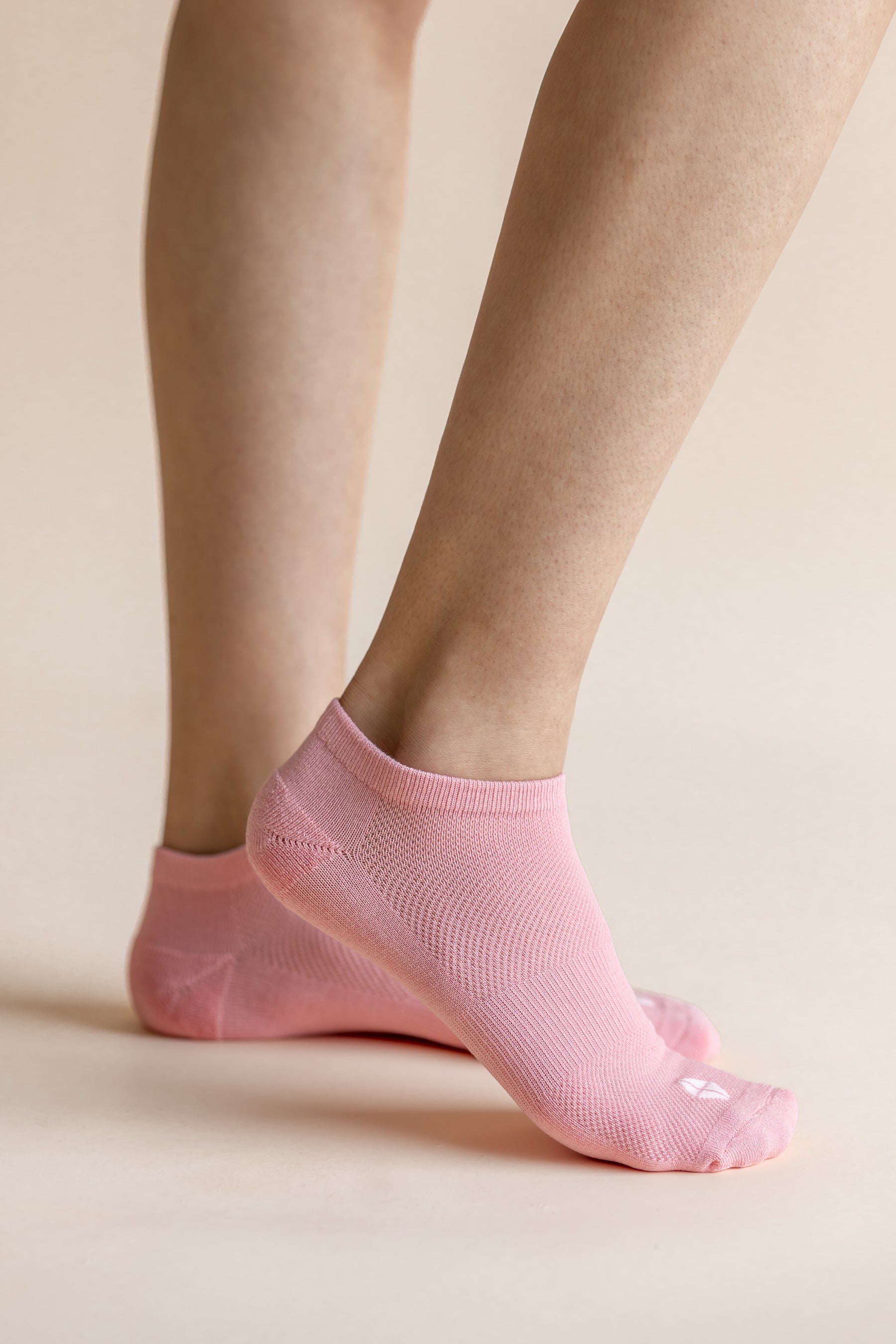 A close-up of a person's feet wearing light pink low-cut ribbed ankle socks against a solid pale beige background.