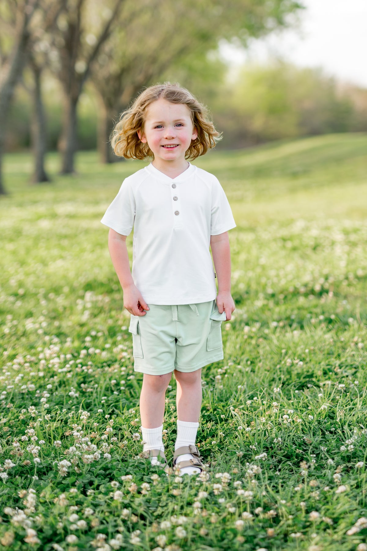 Young child standing in a grass field wearing the Cargo Short in Basil paired with a white short sleeve henley