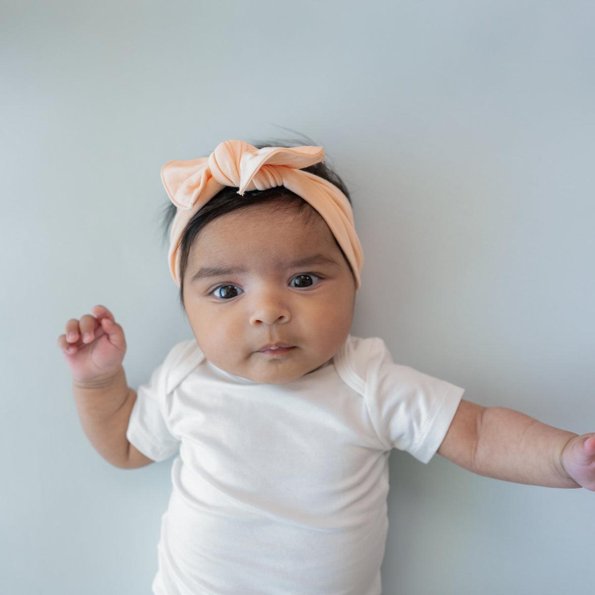 Infant laying down wearing the Knotted Bow Headband in Canyon paired with a Short Sleeve Bodysuit in Cloud