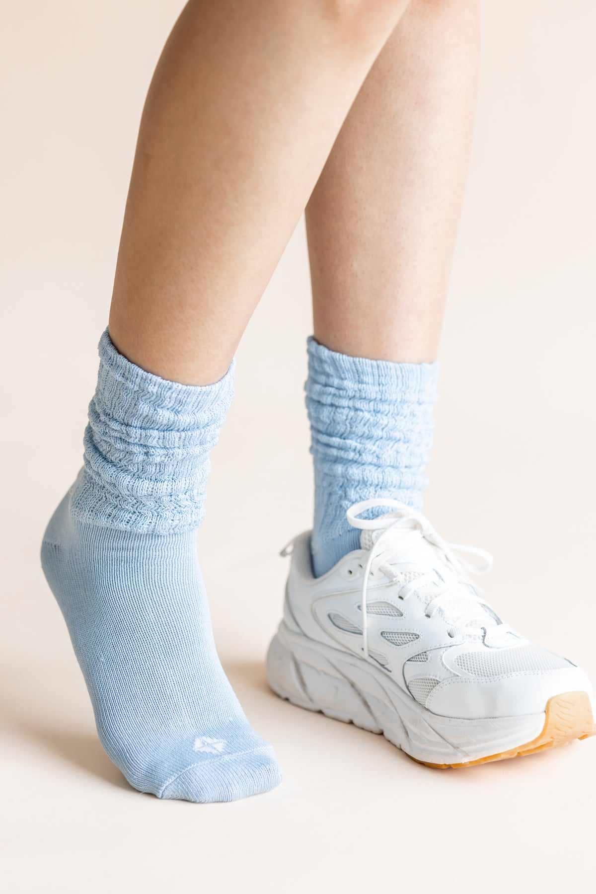 A close-up of a person's legs wearing light blue ribbed crew socks and a white athletic sneaker against a plain beige background.