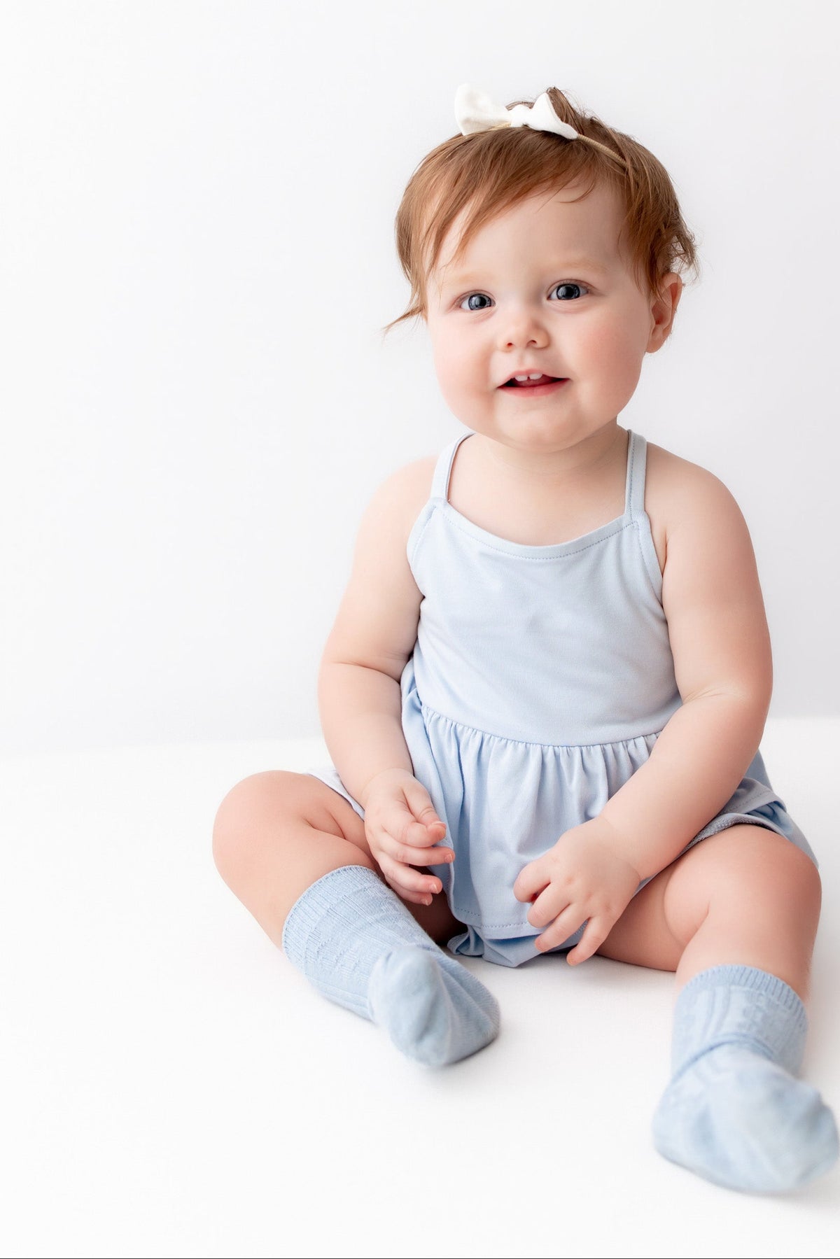 A baby girl sitting on a white studio floor wearing a light blue sleeveless romper and matching socks, with a small white bow in her hair. An adult hand is pointing from the right side.