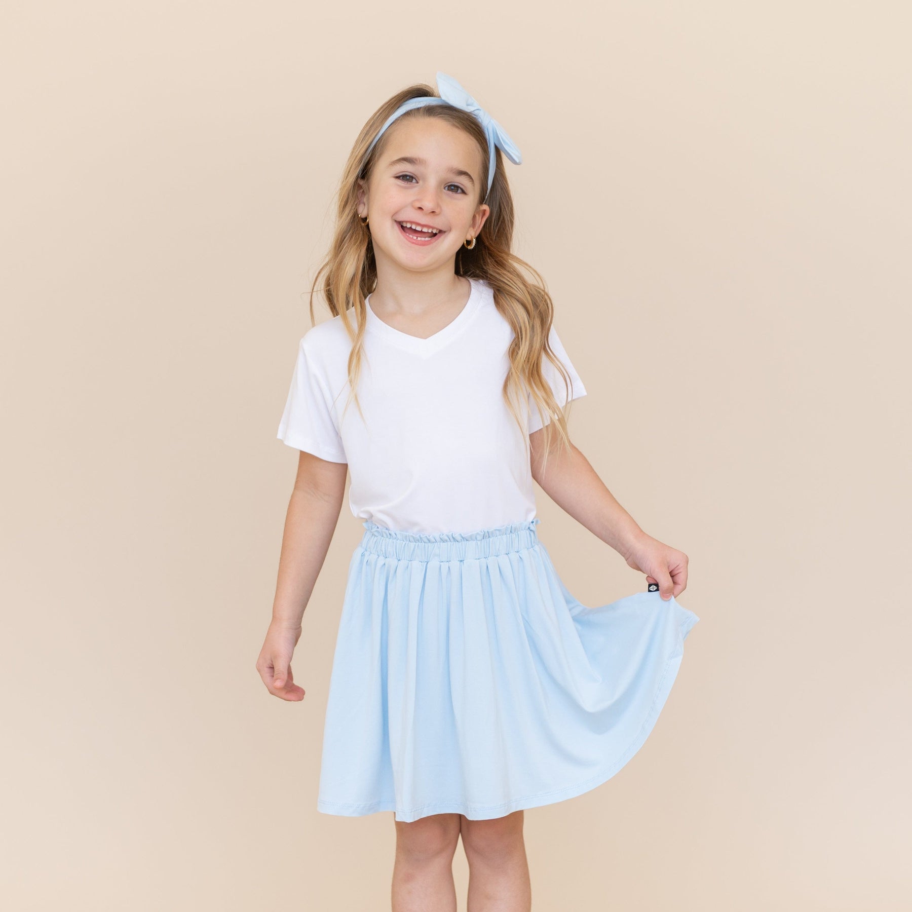 Smiling model wearing the Toddler Skort in Breeze holding the bottom hem out paired with a white v-neck shirt