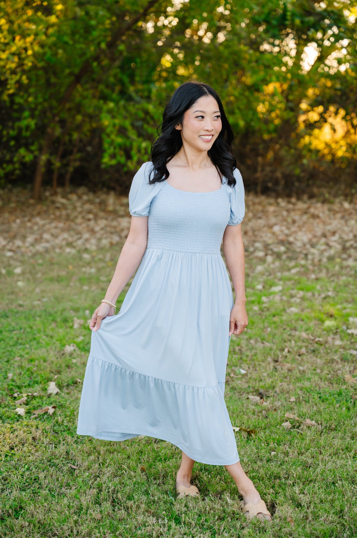 Female modeling the Women’s Smocked Dress in Breeze standing in a grassy field surrounded by trees