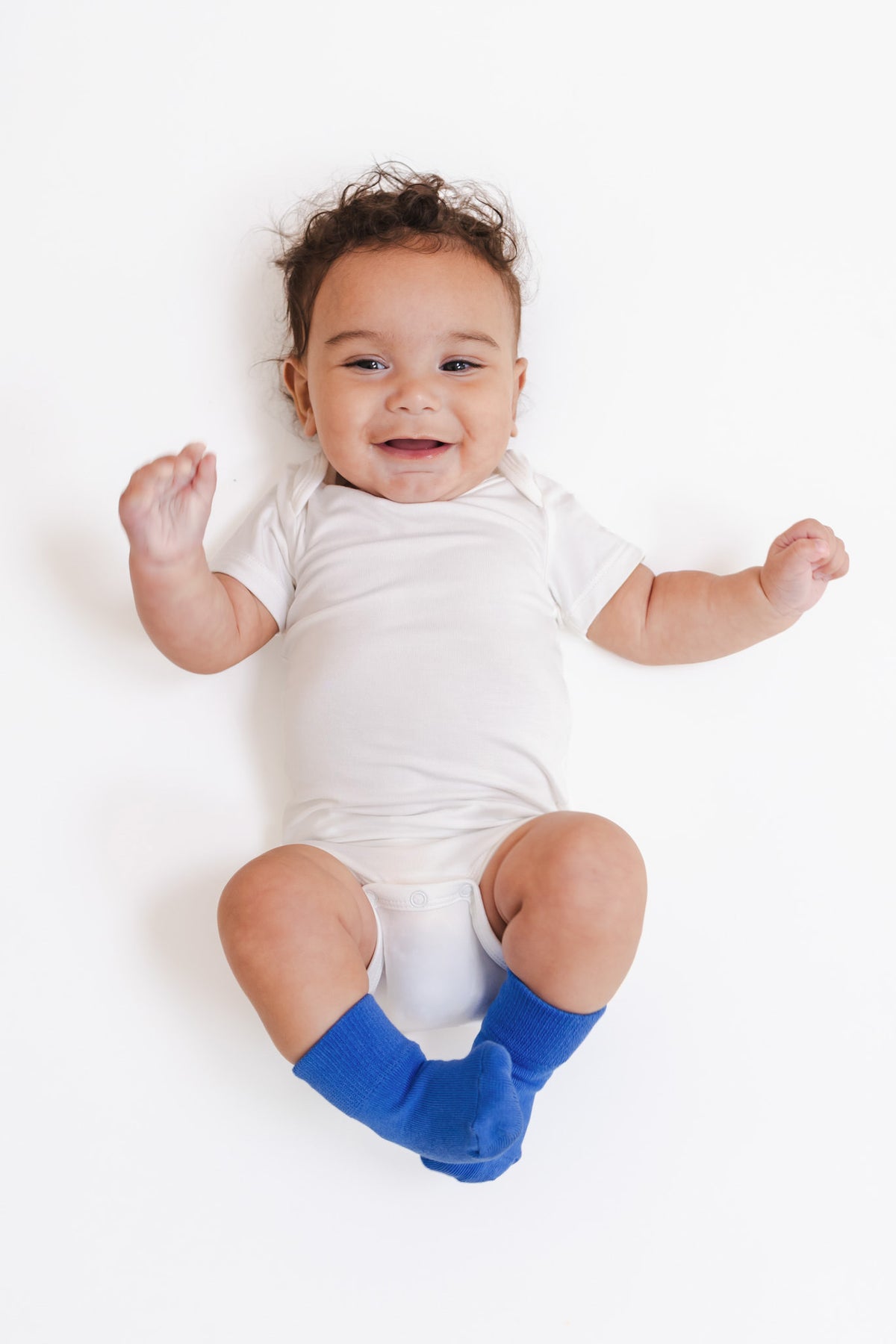 A smiling baby lying on their back wearing a white short-sleeve bodysuit and bright blue socks, photographed against a clean white background.