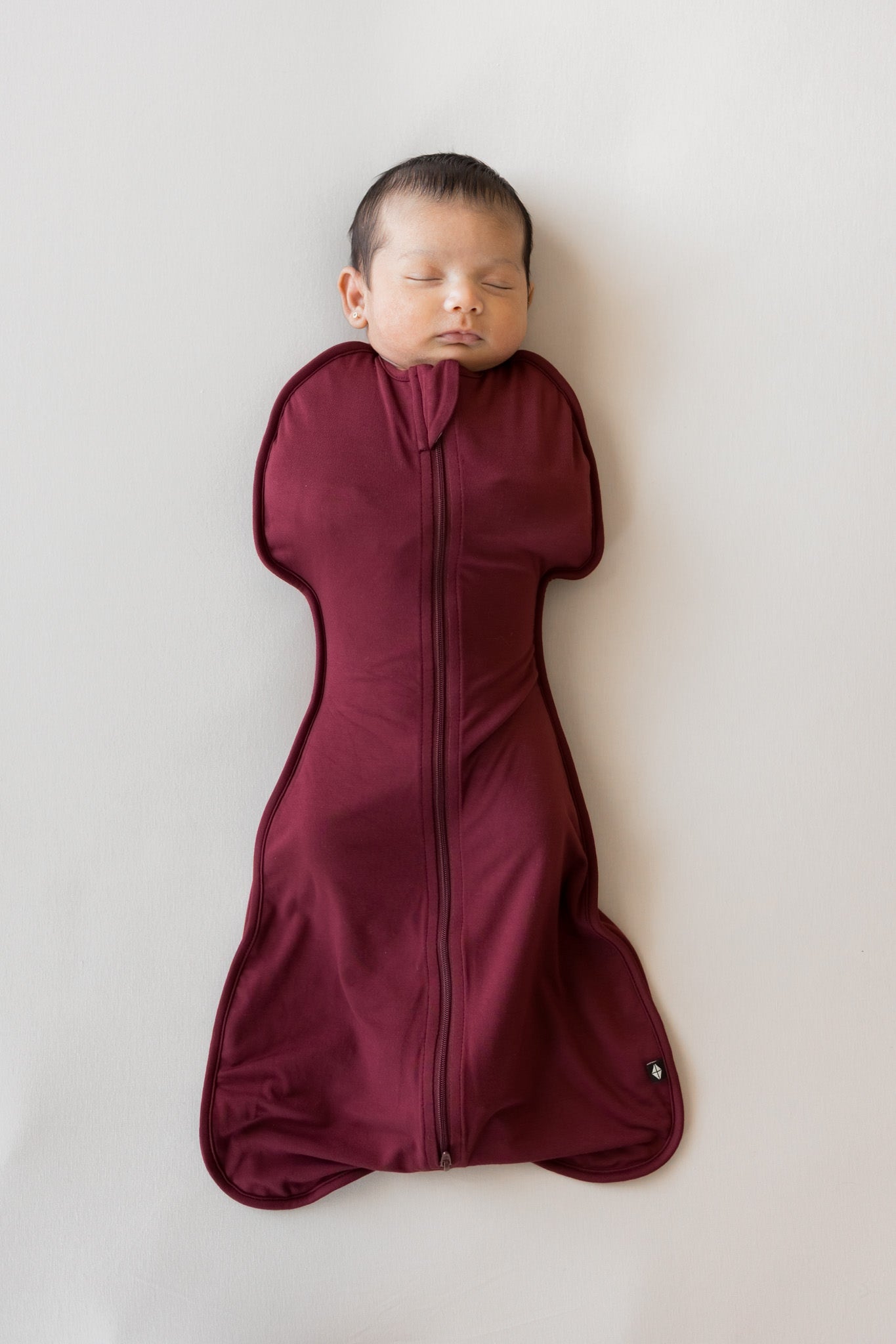 A sleeping newborn baby wrapped in a burgundy zippered swaddle bag, lying flat on a light gray background.