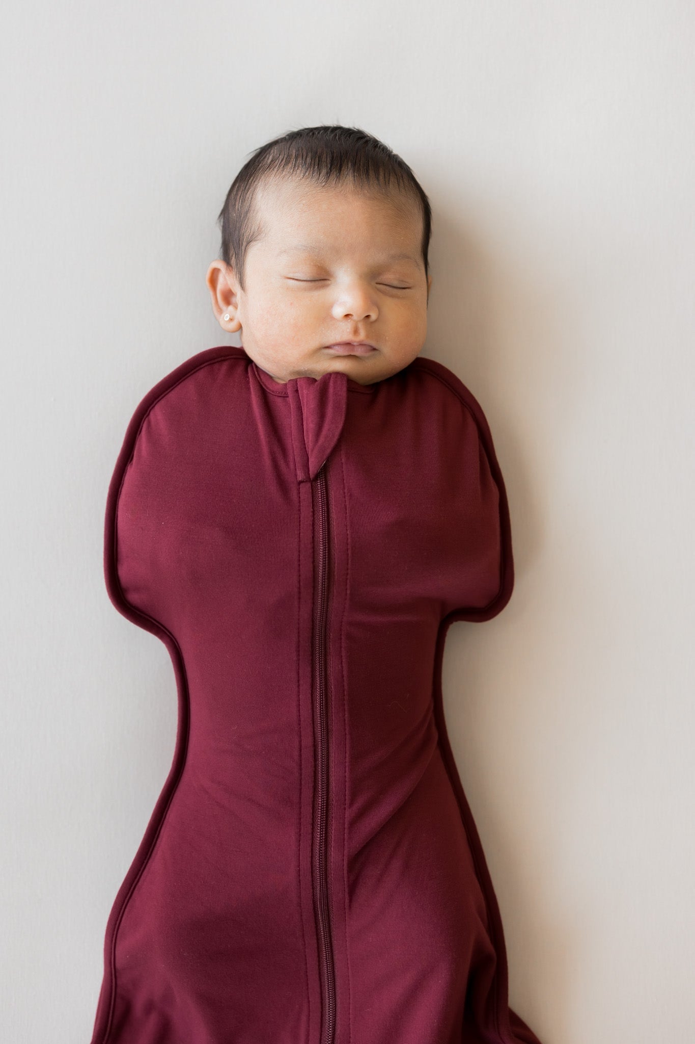 A sleeping newborn baby wrapped in a deep burgundy zippered swaddle bag, lying against a plain light gray background.