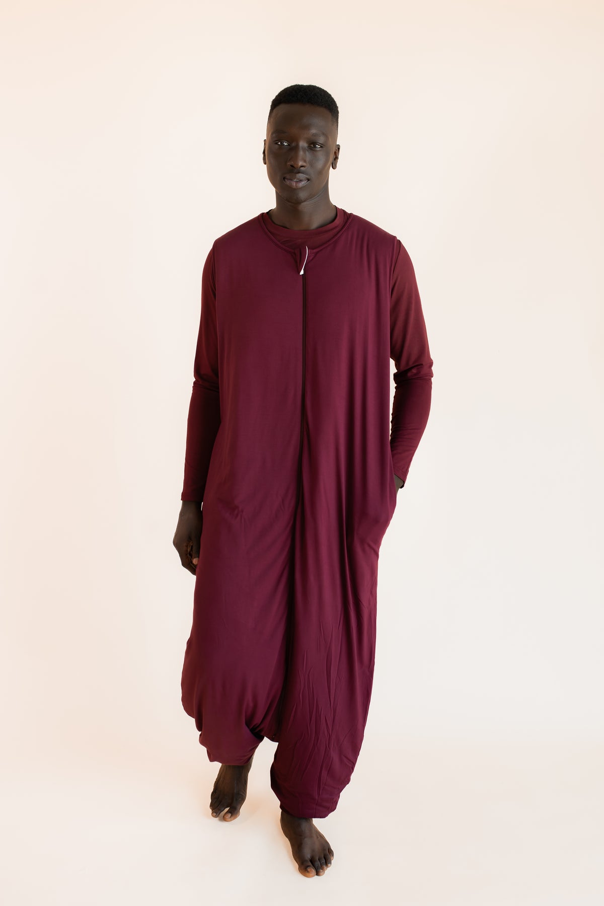 A man wearing a loose-fitting, deep burgundy sleep sack with a front zipper, photographed against a solid light beige background.