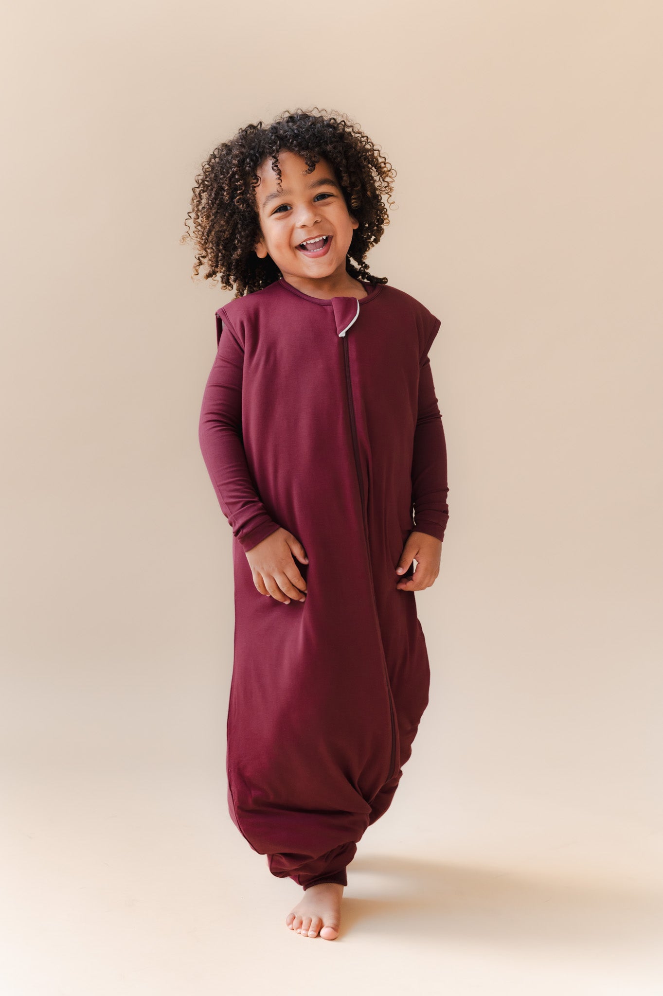 A smiling toddler wearing a burgundy zip-front sleep sack, standing barefoot against a plain beige studio background.