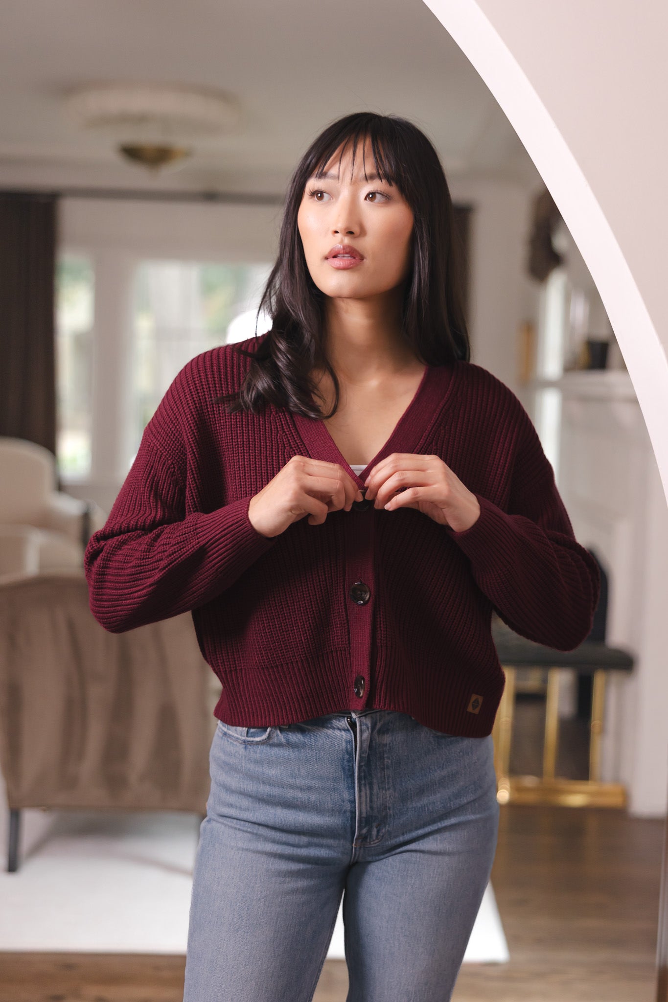 Modal buttoning the Chunky Knit Women's Cropped Cardigan in Burgundy wearing over blue jeans