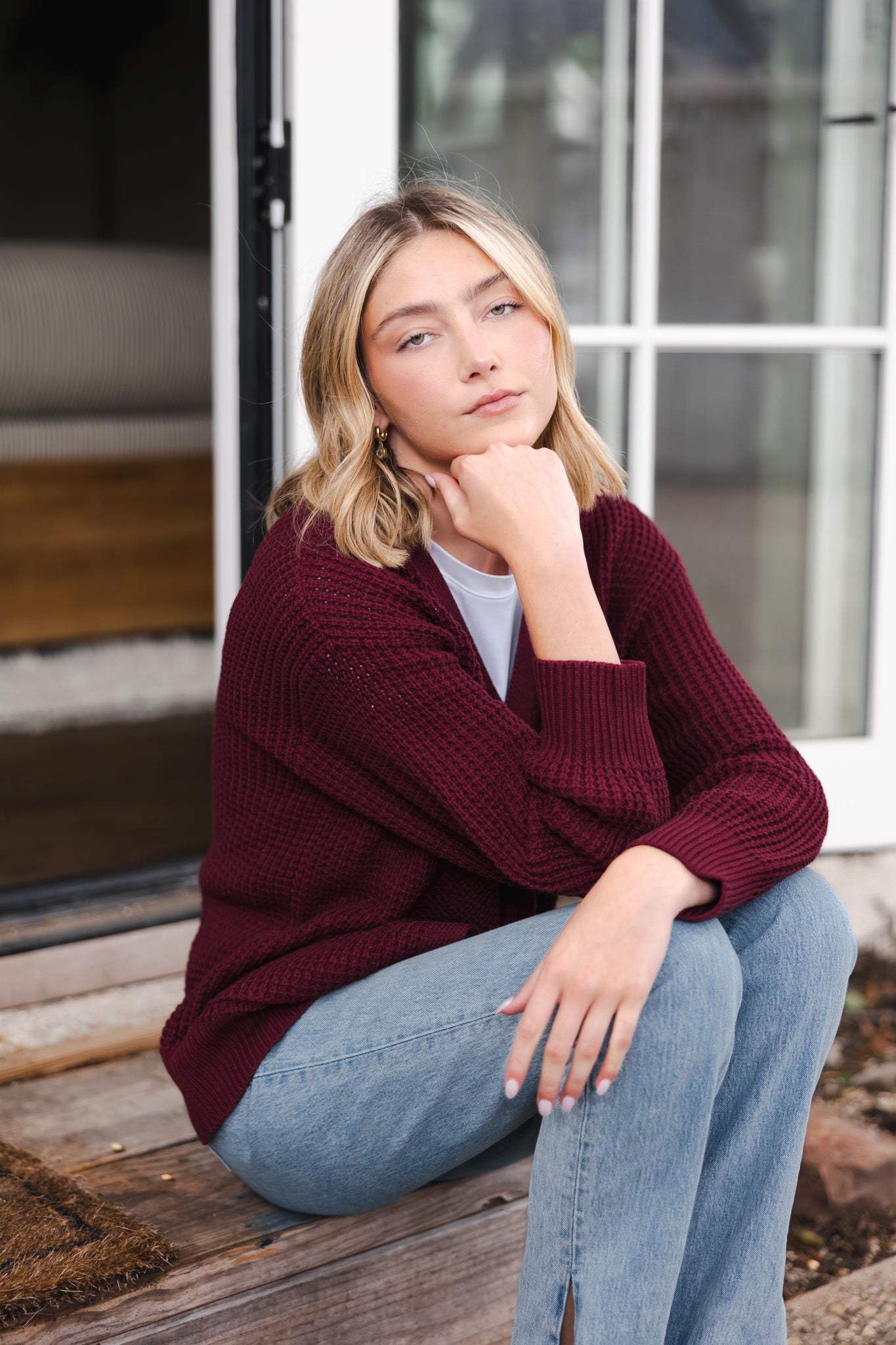 Modal posing outdoors wearing Chunky Knit Women's Waffle Cardigan in Burgundy over white T-shirt and blue jeans