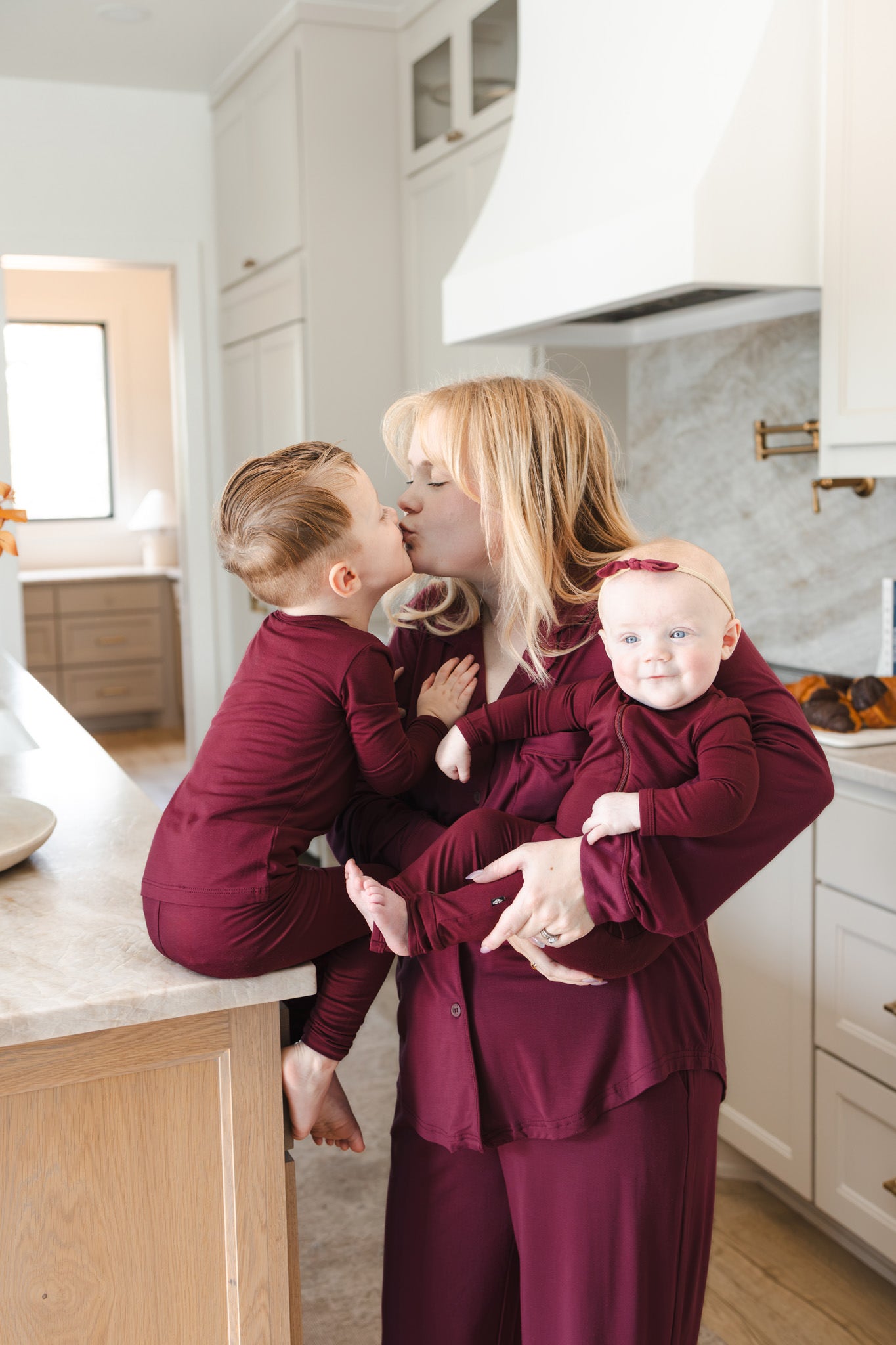 A woman and two children wearing matching burgundy long-sleeve pajamas pose together in a bright, modern kitchen. The woman is holding a baby on her hip while a toddler sits on the kitchen island, all sharing a sweet moment.