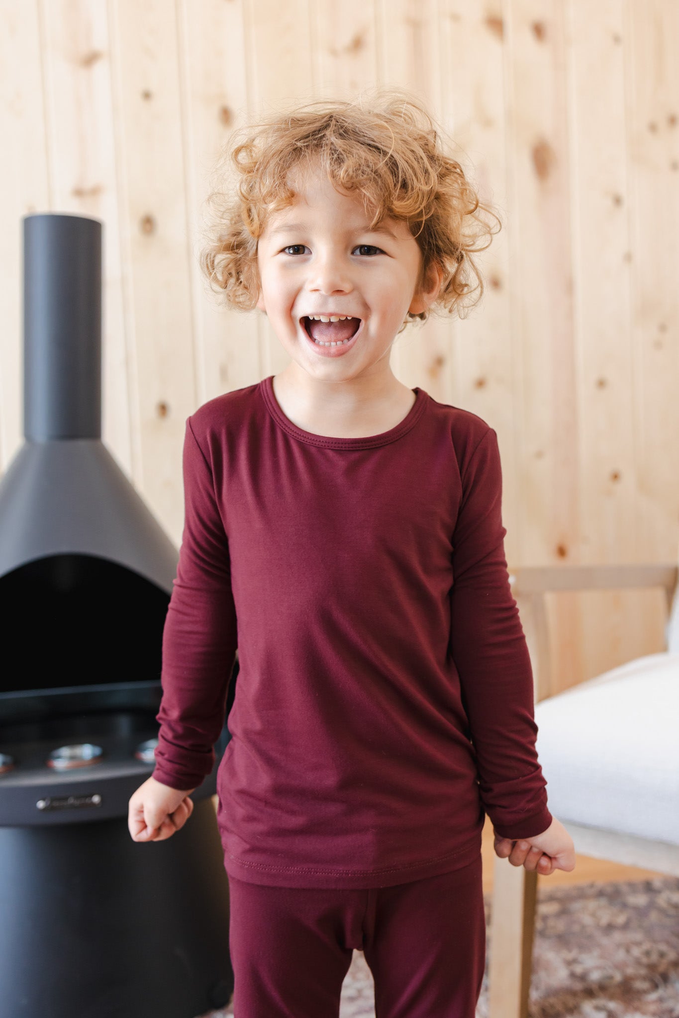 A young child wearing a matching burgundy long-sleeve top and pants set, standing in a room with a wood-paneled wall and a black fireplace.