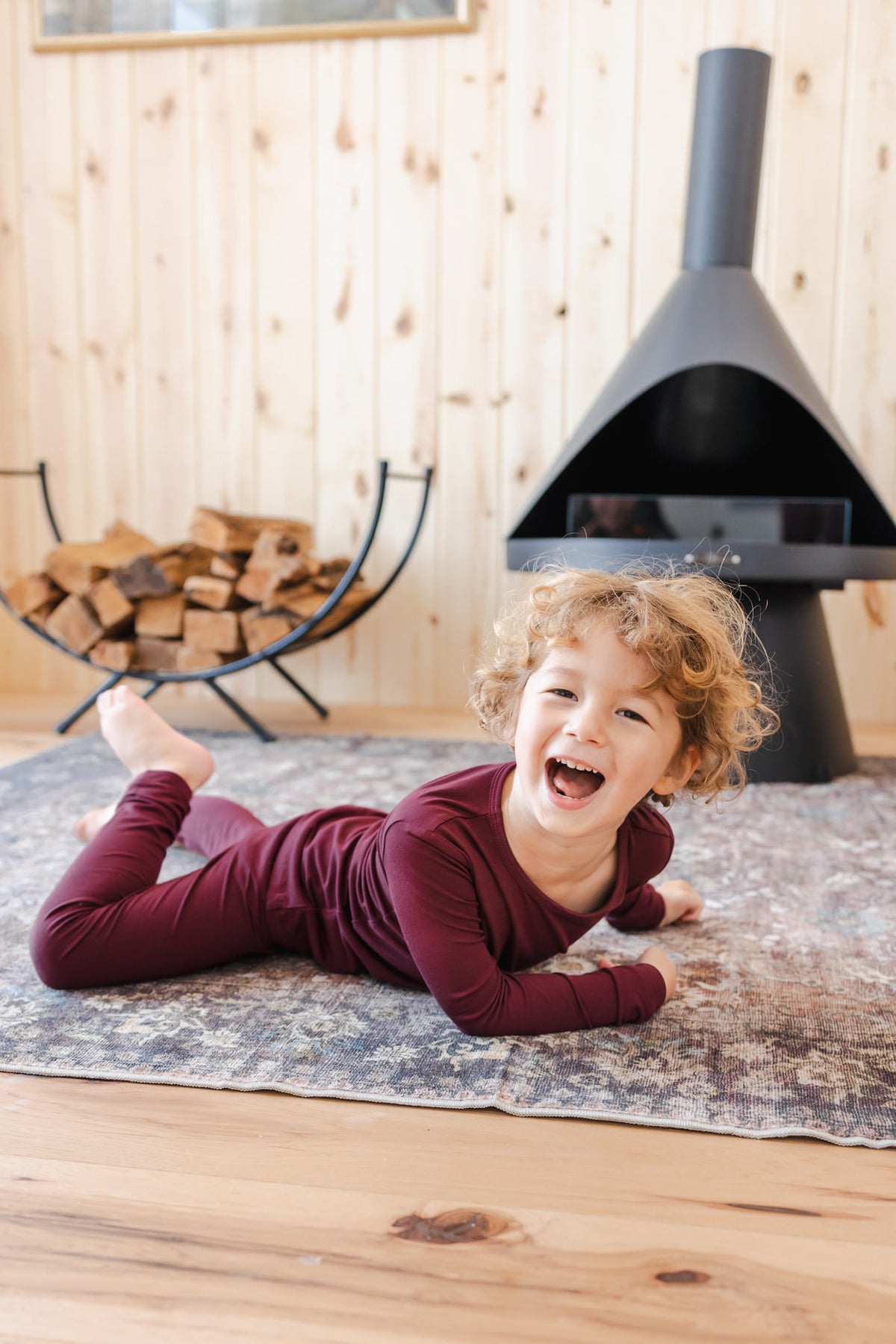 A young child wearing a matching burgundy long-sleeve top and pant pajama set. A black fireplace and a wood stack are visible in the background.