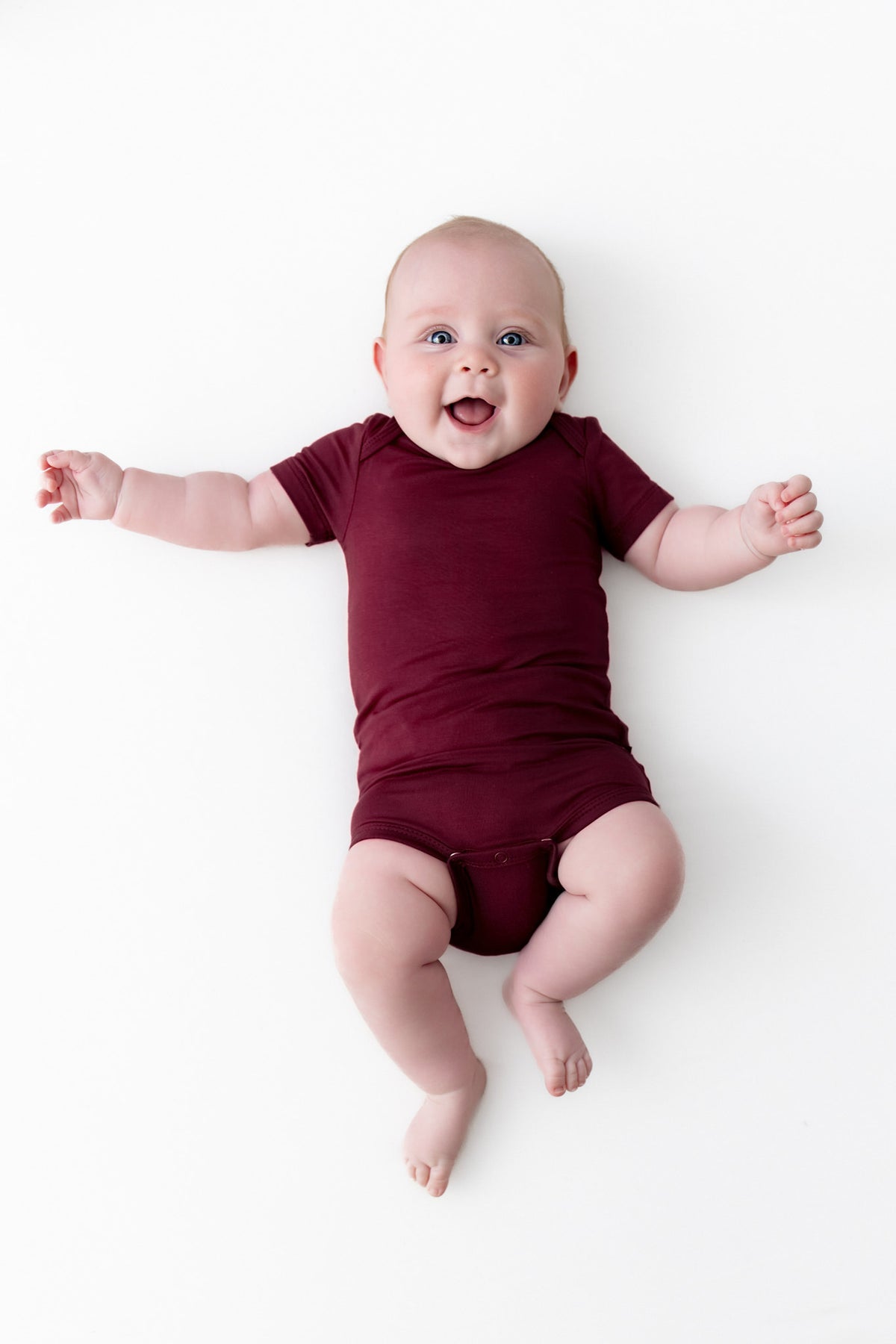 A smiling baby lying on their back wearing a short-sleeve burgundy baby bodysuit against a solid white background.