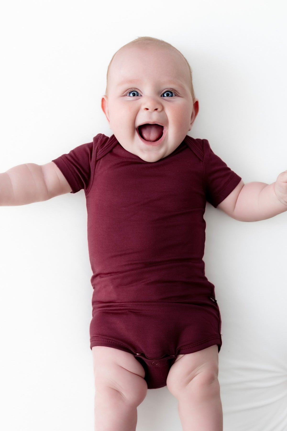 A baby lying on a white surface wearing a short-sleeve burgundy onesie