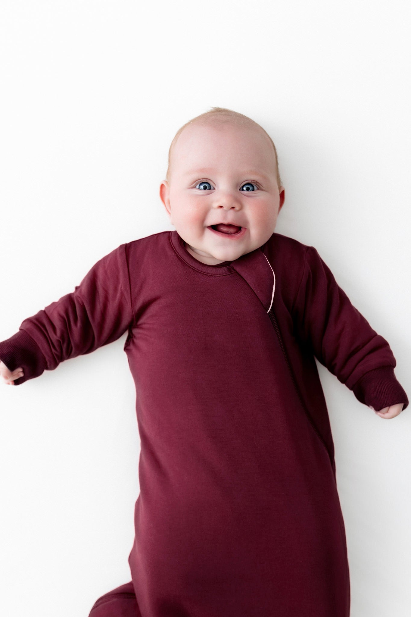 A smiling baby lying on their back wearing a long-sleeve burgundy sleep sack against a plain white background.