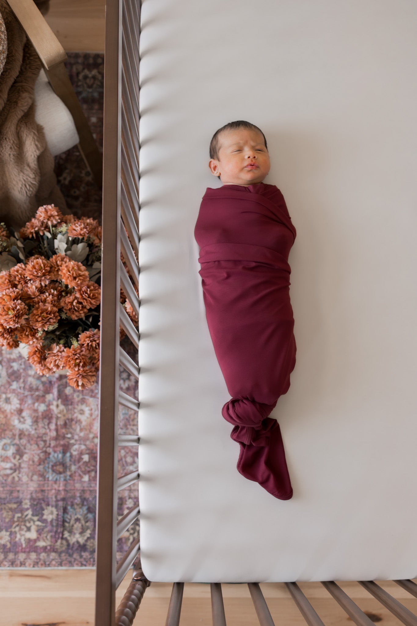 A sleeping newborn baby swaddled in a deep burgundy wrap blanket, lying on a white mattress inside a wooden crib.