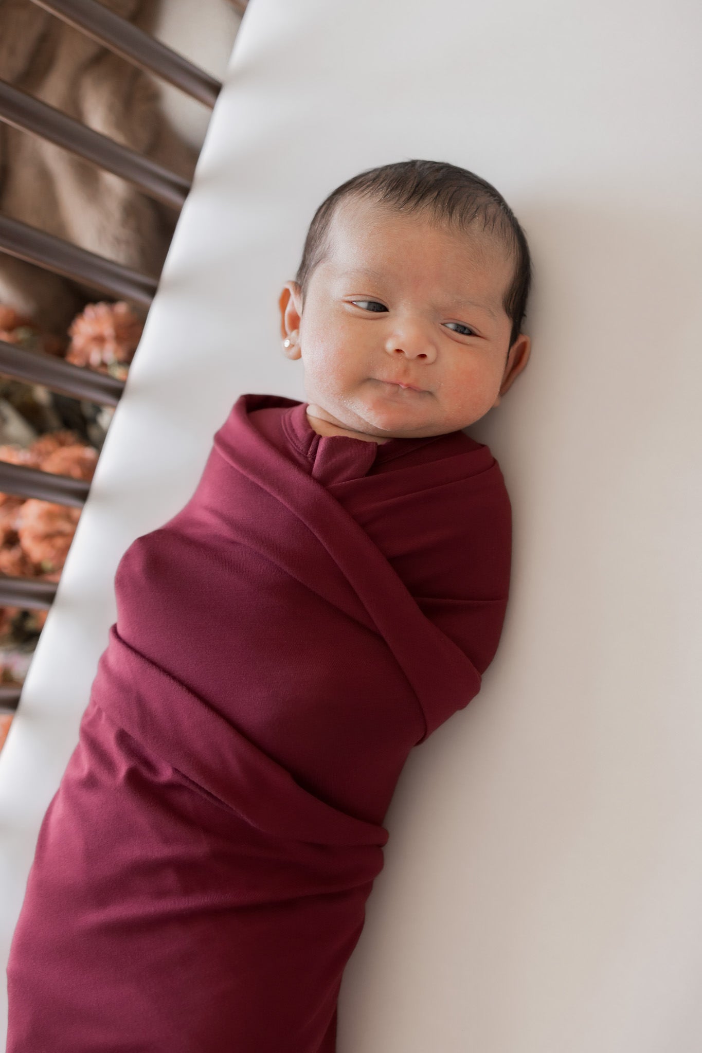 A newborn baby swaddled in a deep burgundy wrap blanket, lying on a white blanket next to a crib.