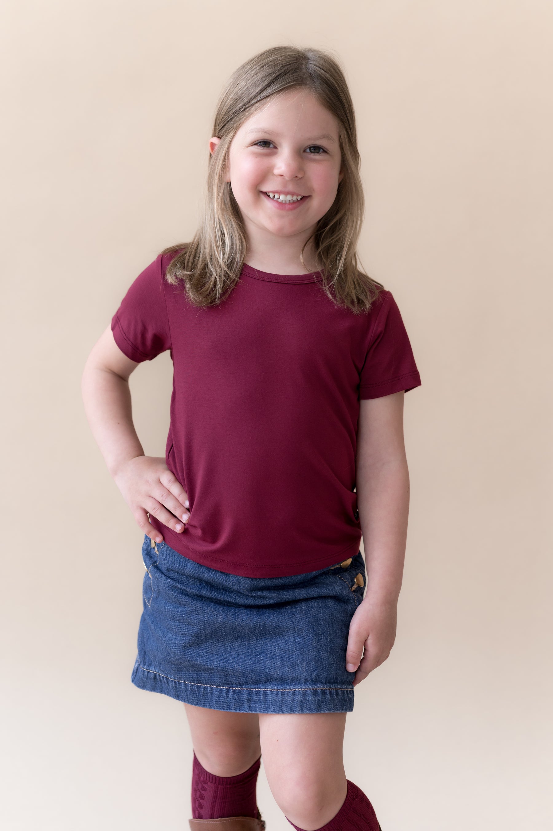 Girl wearing Toddler Basic Tee in Burgundy on blue skirt and burgundy knee high socks and boots on a beige background