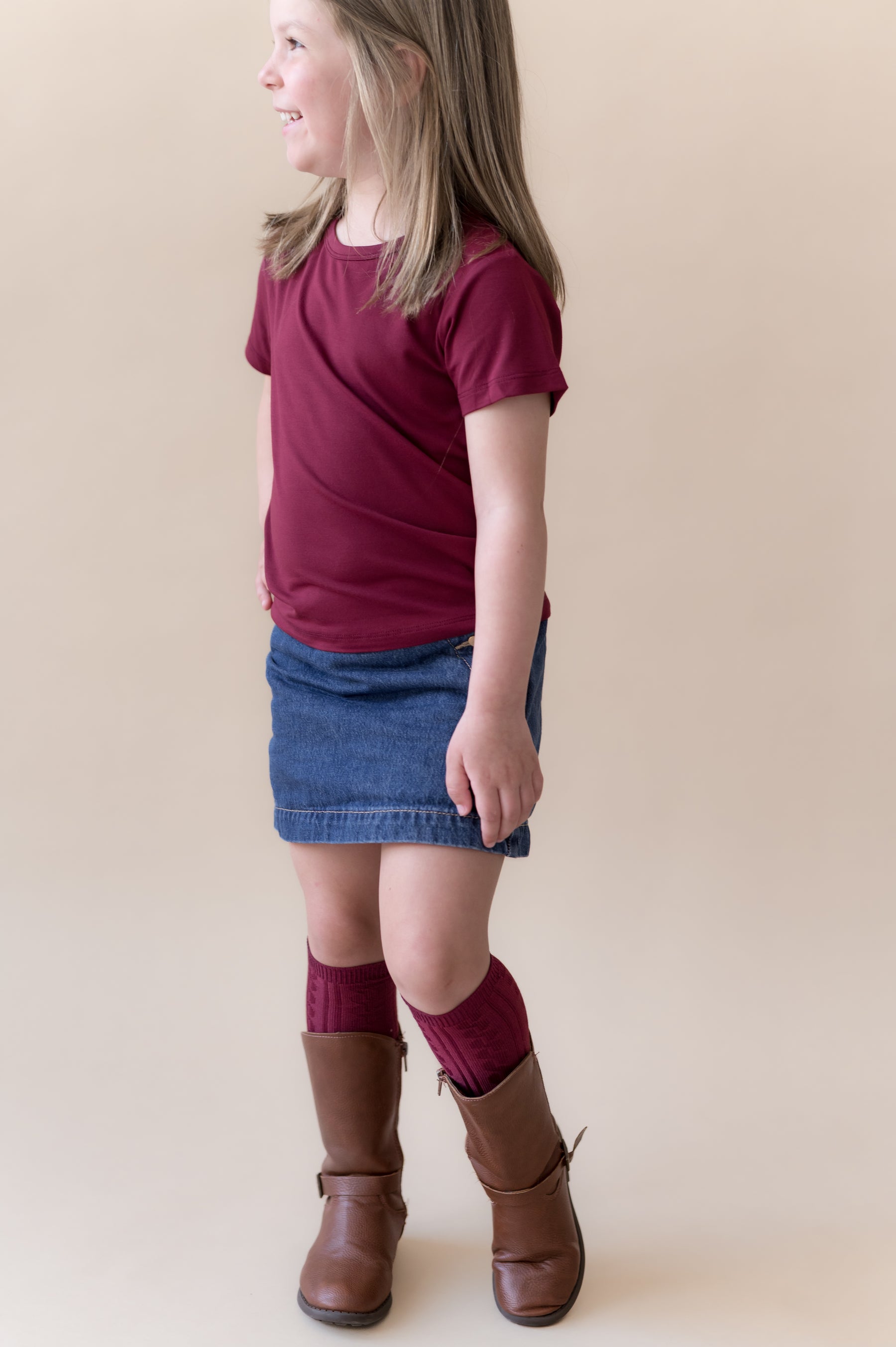 Girl wearing Toddler Basic Tee in Burgundy on blue skirt and burgundy knee high socks and boots on a beige background
