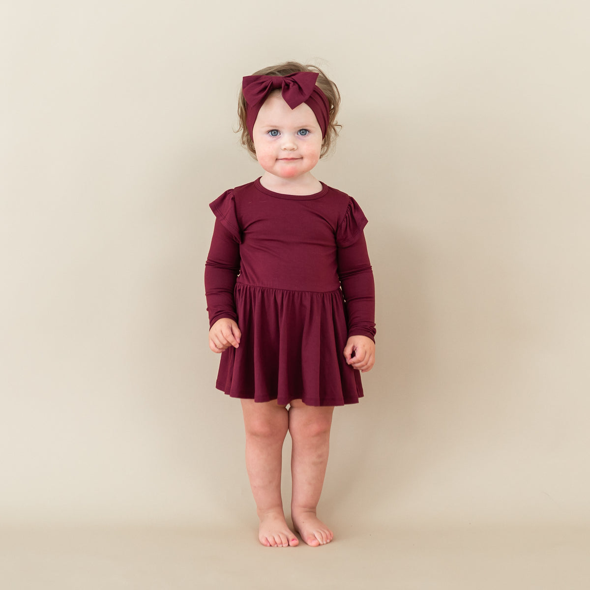 Toddler standing wearing the Long Sleeve Twirl Bodysuit Dress in Burgundy with matching bow headband