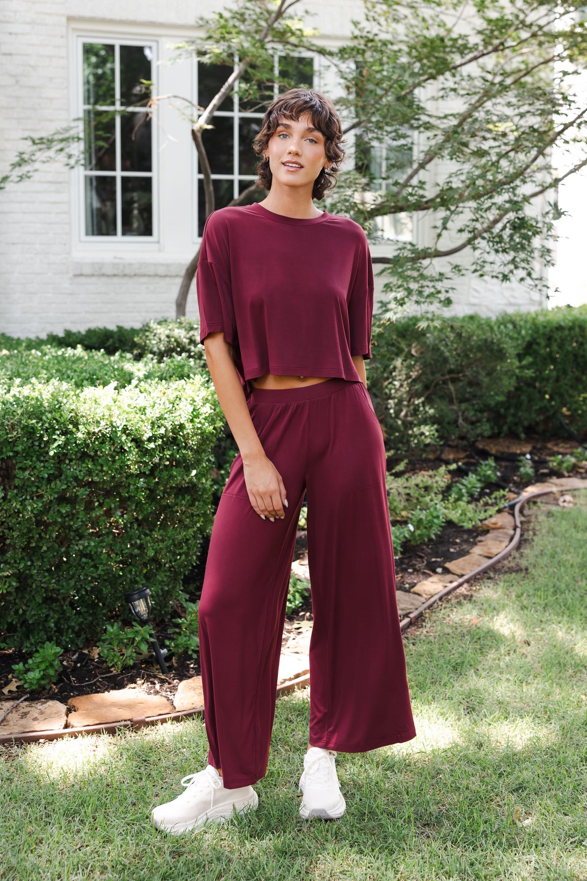 Female model standing in garden wearing the Women's Wide Leg Pant in Burgundy and matching short sleeve tee on white shoes