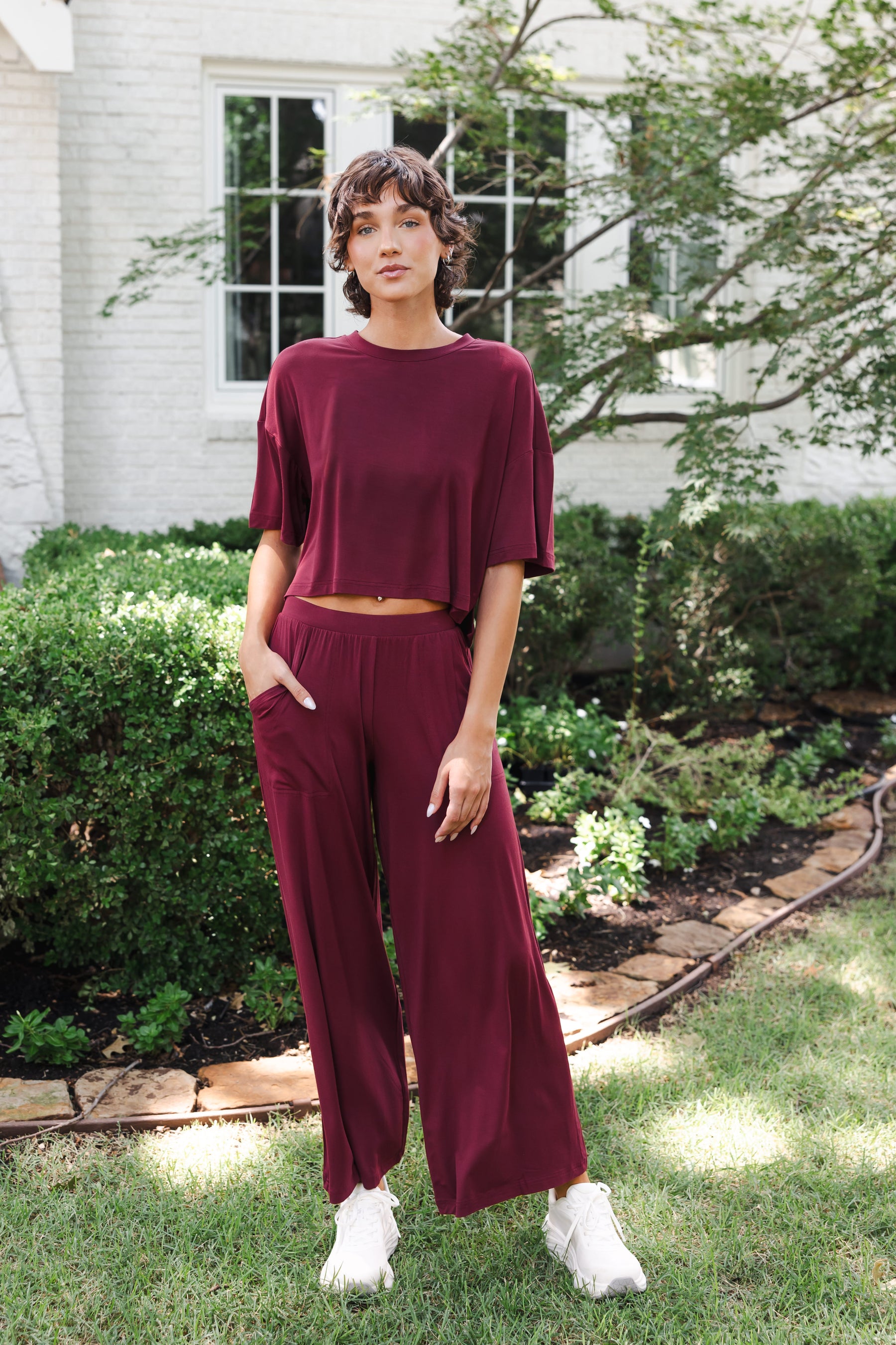 Female model standing in garden wearing the Women's Wide Leg Pant in Burgundy with pockets and matching short sleeve tee on white shoes