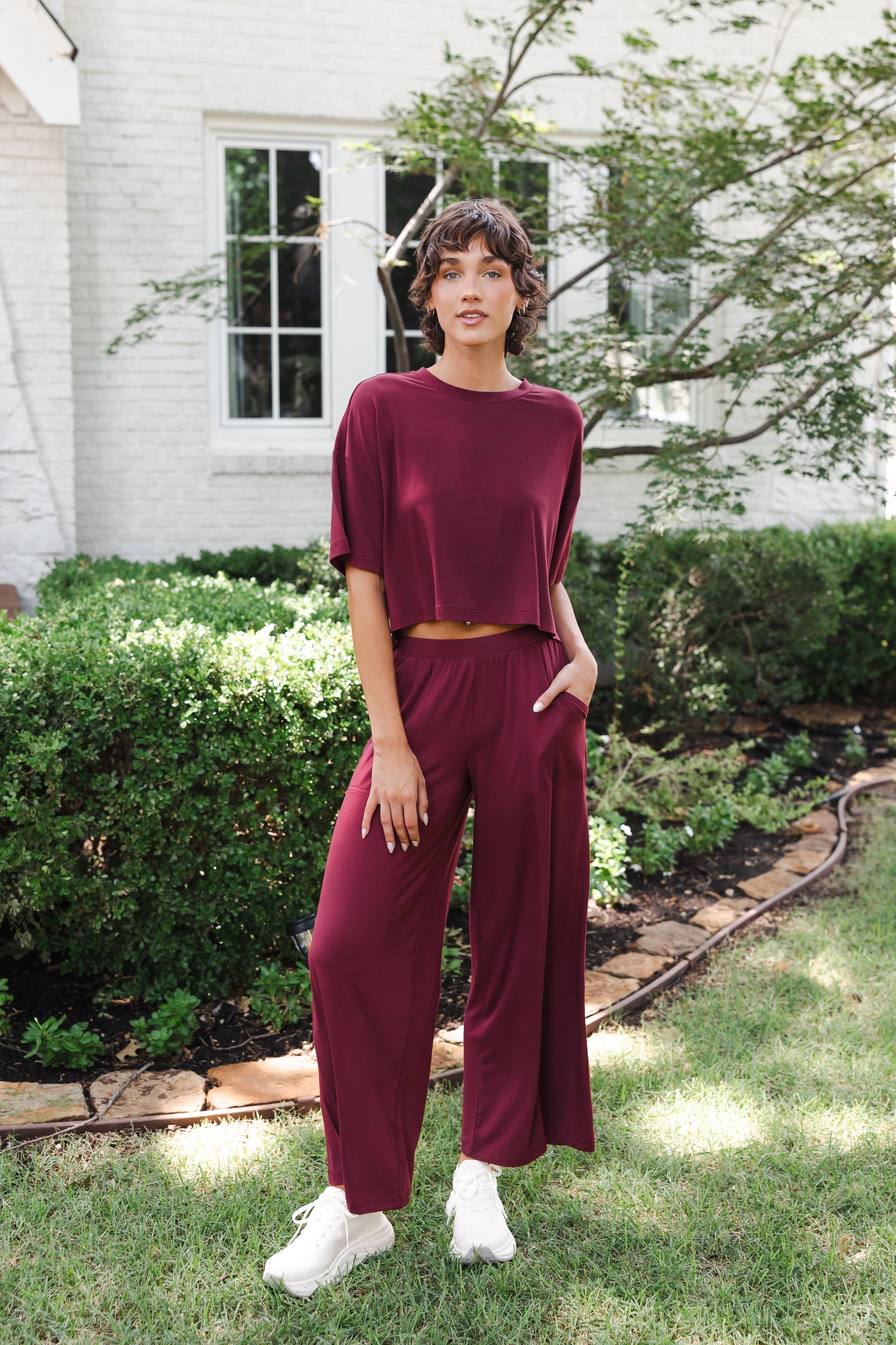 Female model posing in garden wearing the Women's Wide Leg Pant in Burgundy and matching short sleeve tee on white shoes