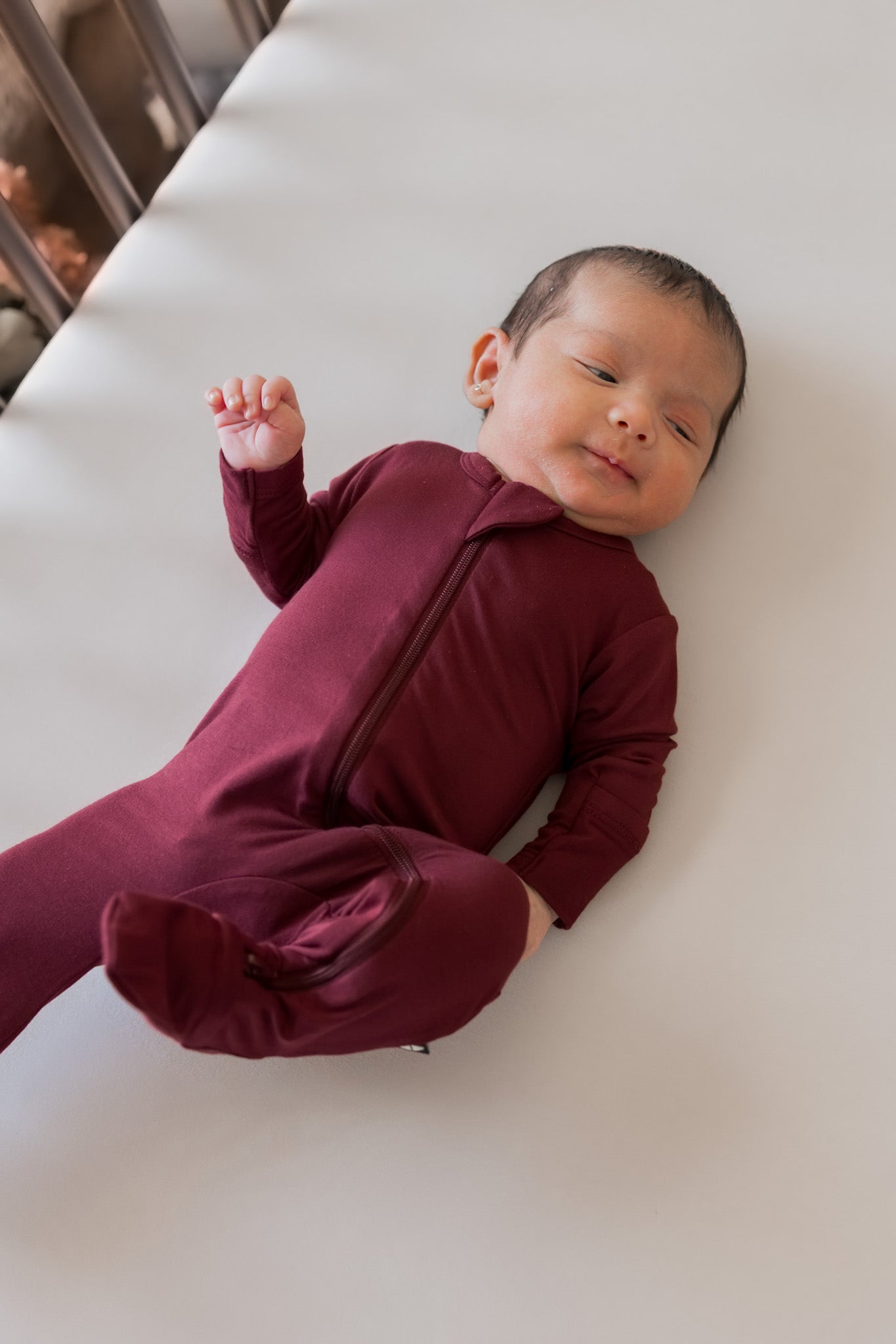 A newborn baby wearing a burgundy long-sleeve footless sleeper lies on their back on a white surface.