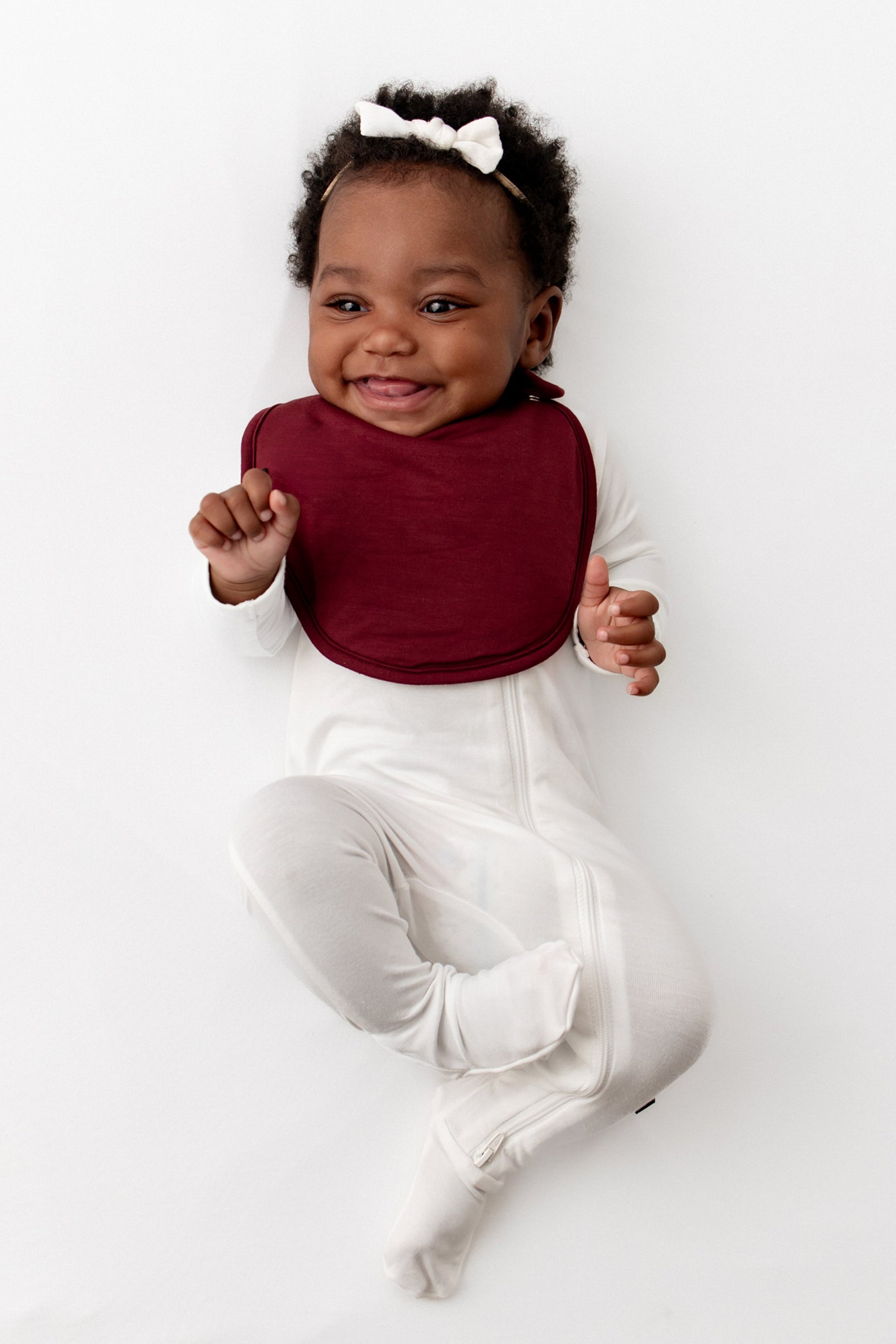 Smiling infant wearing the Cushy Bib in Burgundy overtop a Cloud zippered footie