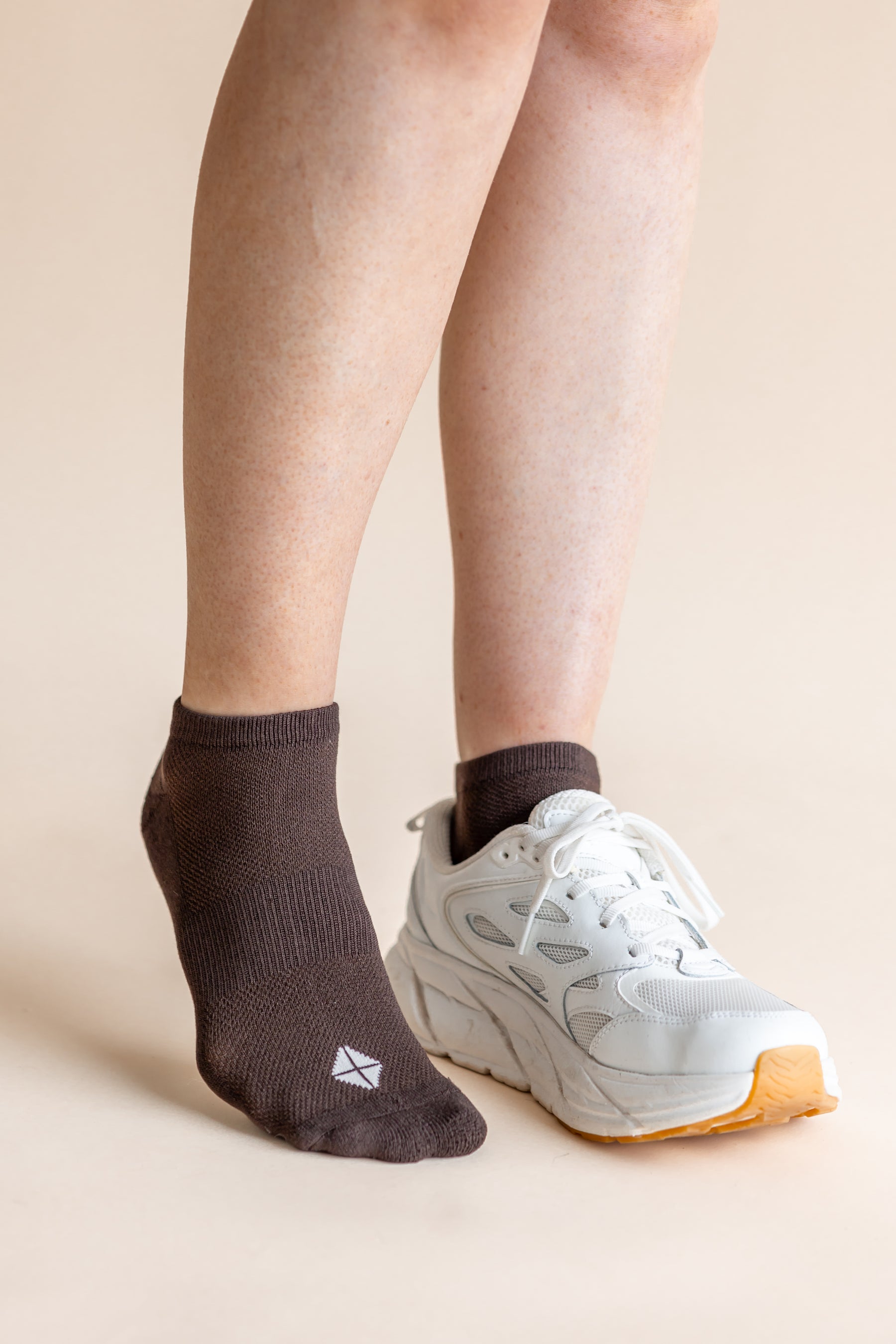 A person's legs and feet shown wearing a dark brown ankle sock on one foot and a white athletic sneaker on the other, against a plain beige studio background.