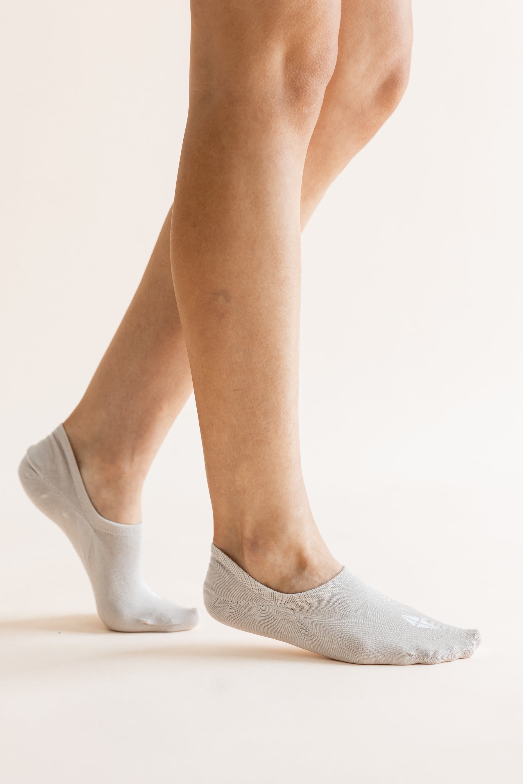 A close-up of a person's feet wearing light gray low-cut no-show socks against a plain, warm off-white background.
