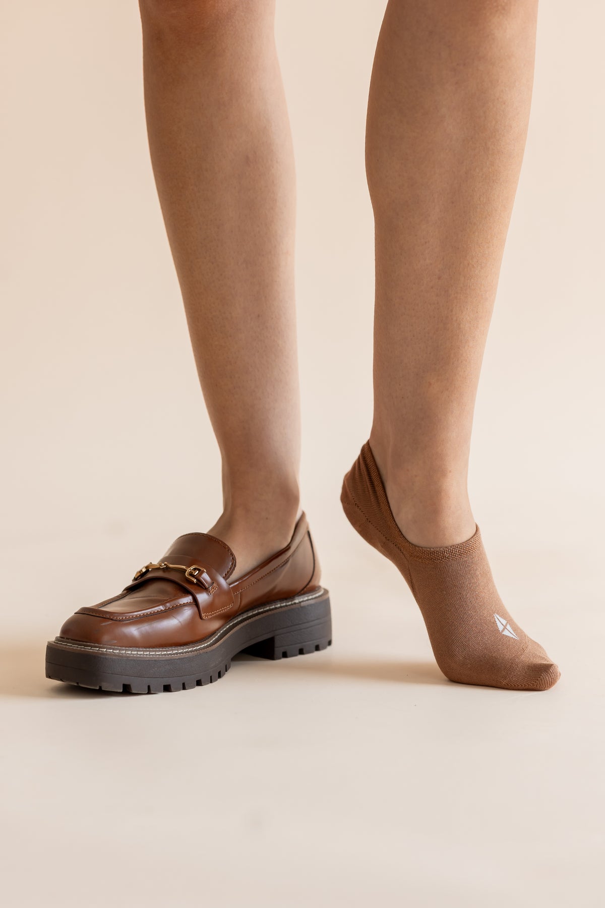 A person wearing brown leather loafers with chunky black platform soles and tan ankle socks, shown from the calves down against a plain beige studio background.