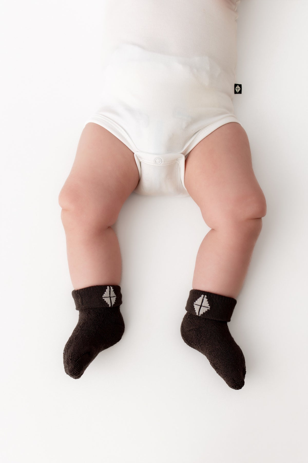 A baby lying on a white surface wearing white bodysuit bottoms and dark brown socks with a small white geometric logo near the cuff.