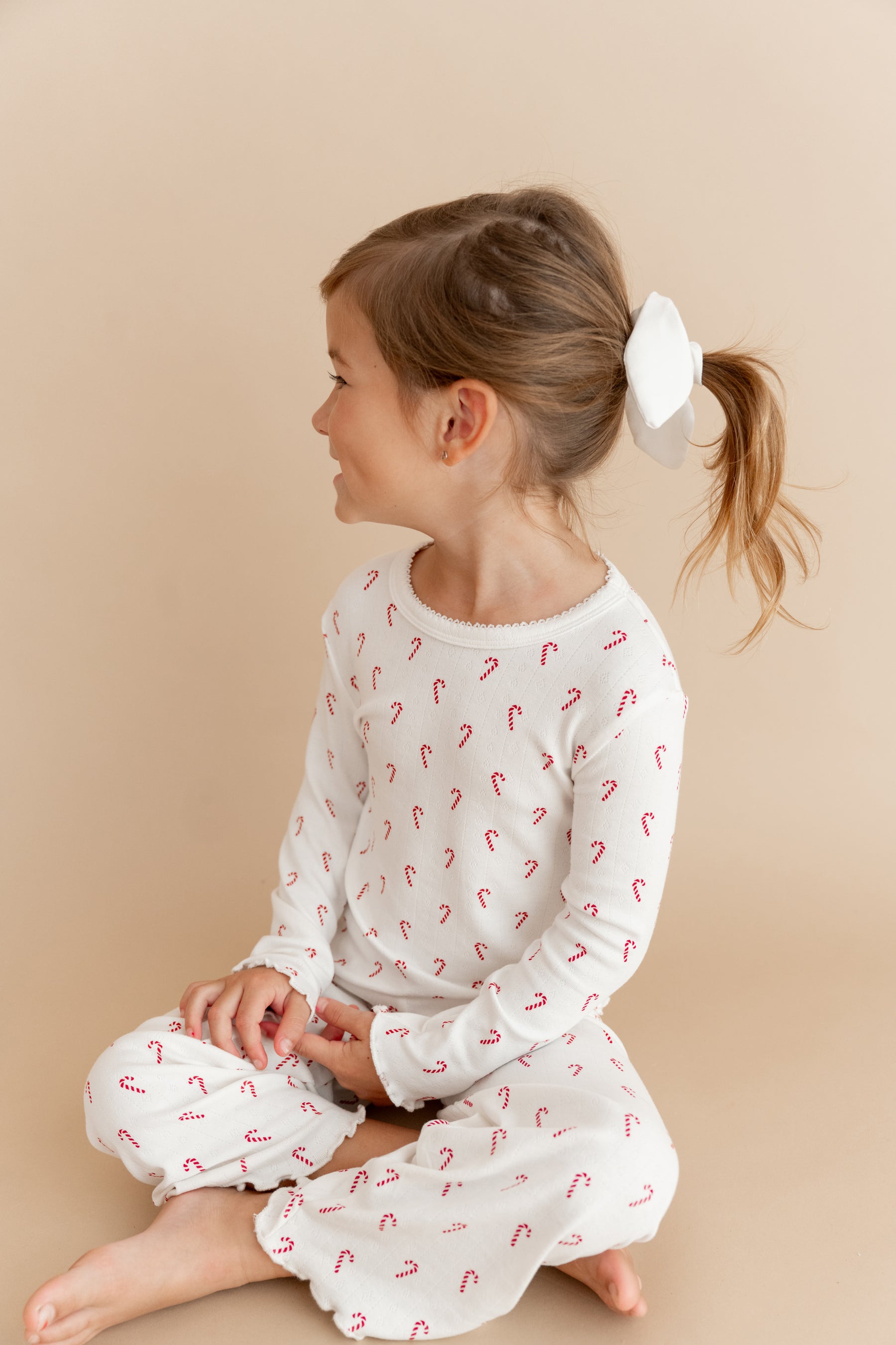 A young girl with her hair in a ponytail sits cross-legged on a plain beige studio background, wearing a white long-sleeve pointelle set with a small red candy cane print.