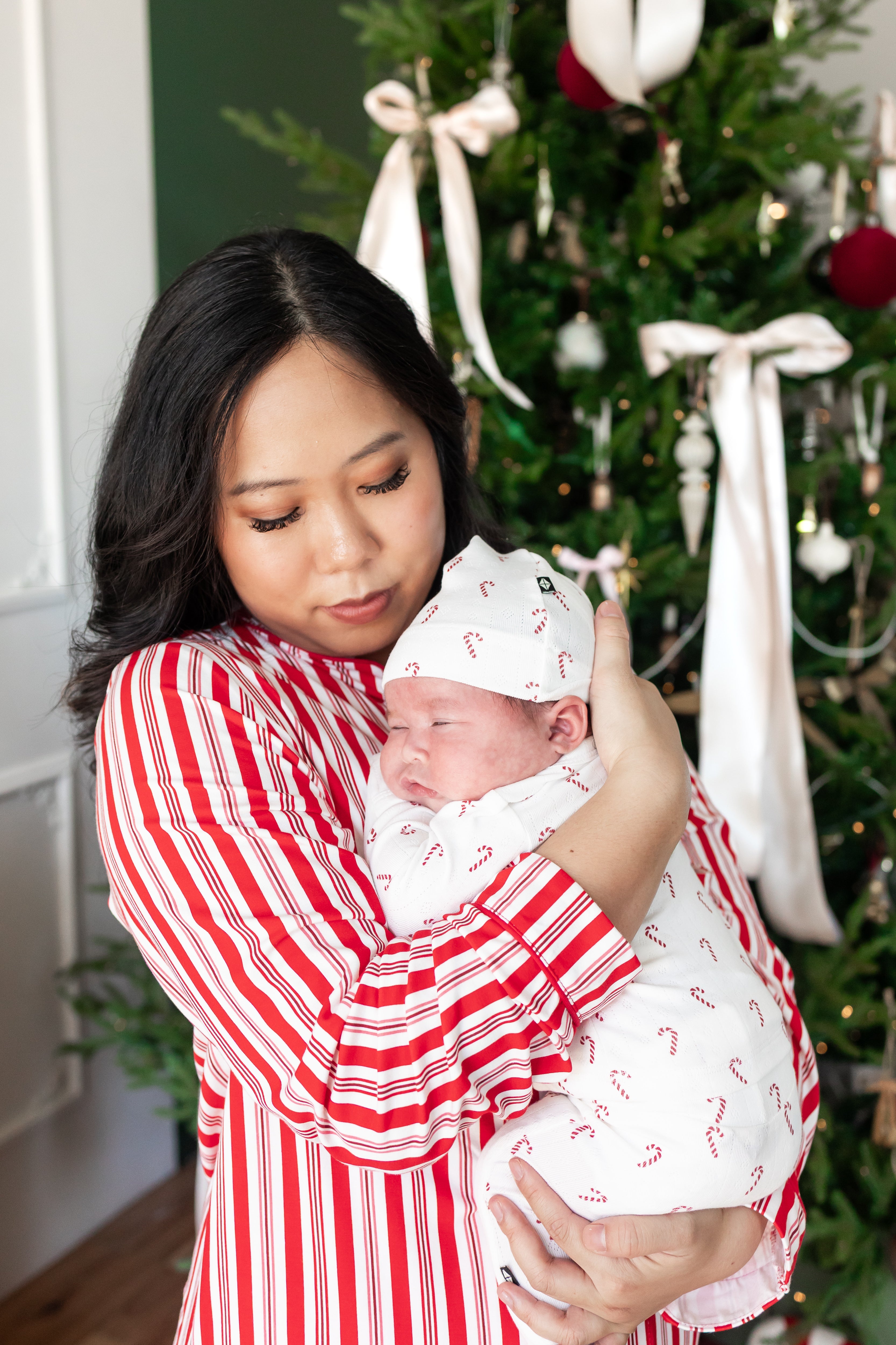Mommy wearing striped red and white long sleeve pajama set holding her sleeping baby against her chest wearing Pointelle Take Me Home Set with Cap in Candy Cane print near a Christmas tree