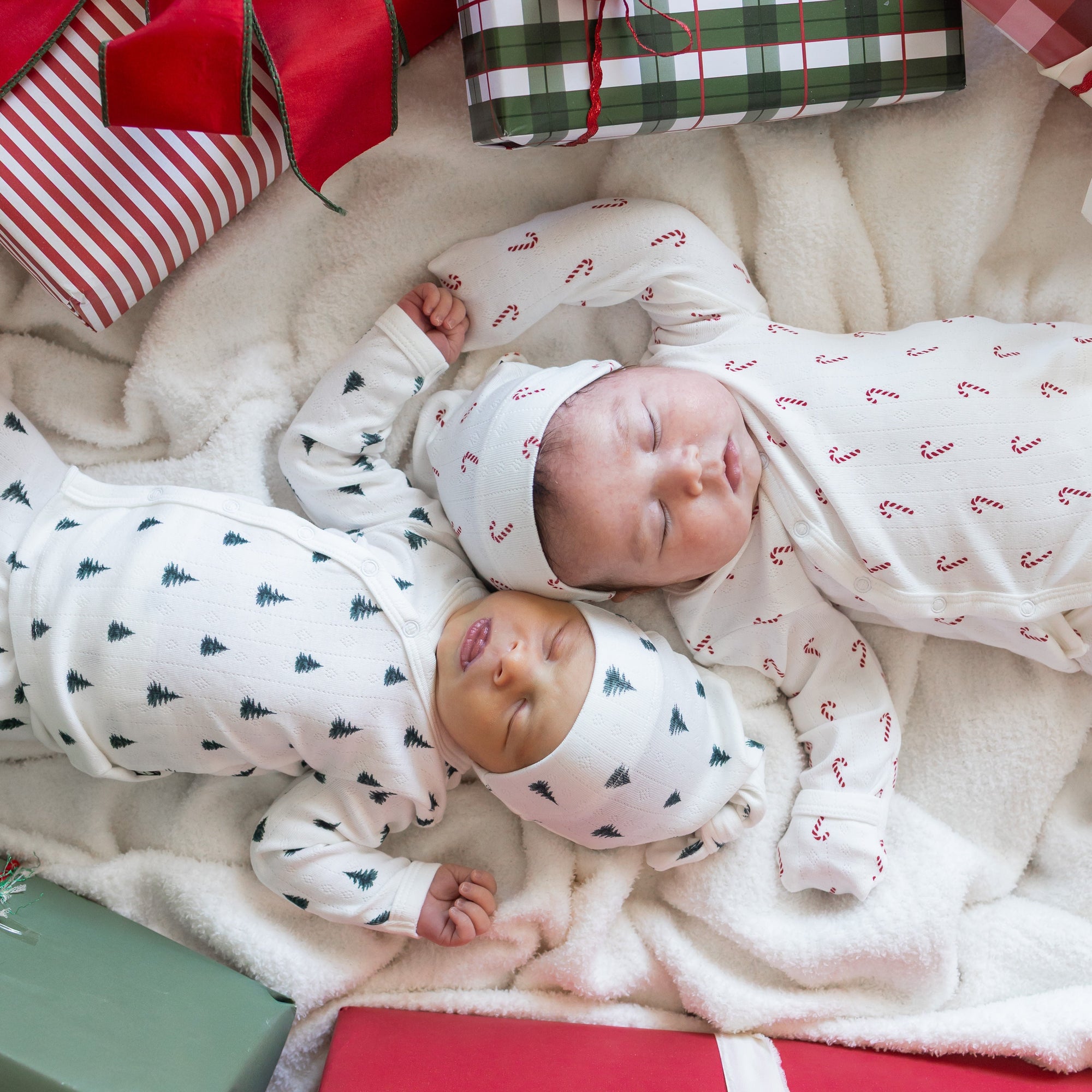 2 newborn babies sleeping wearing pointelle take me home sets in delicate christmas patterns laying near christmas presents 