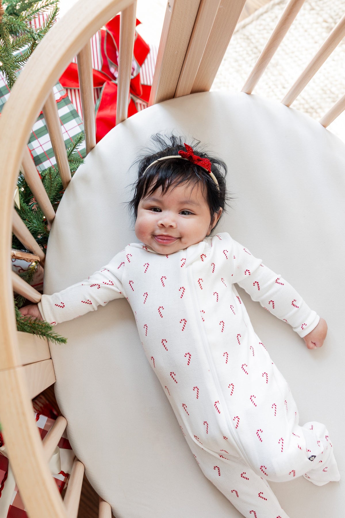 Baby wearing Pointelle Zippered Footie in Candy Cane with a headband lying in a crib near a Christmas tree
